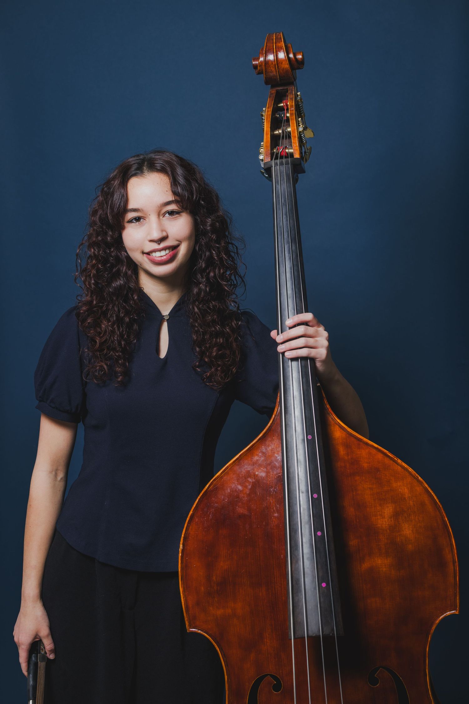 Young female double bass player with curly hair holding her instrument against a dark blue background.