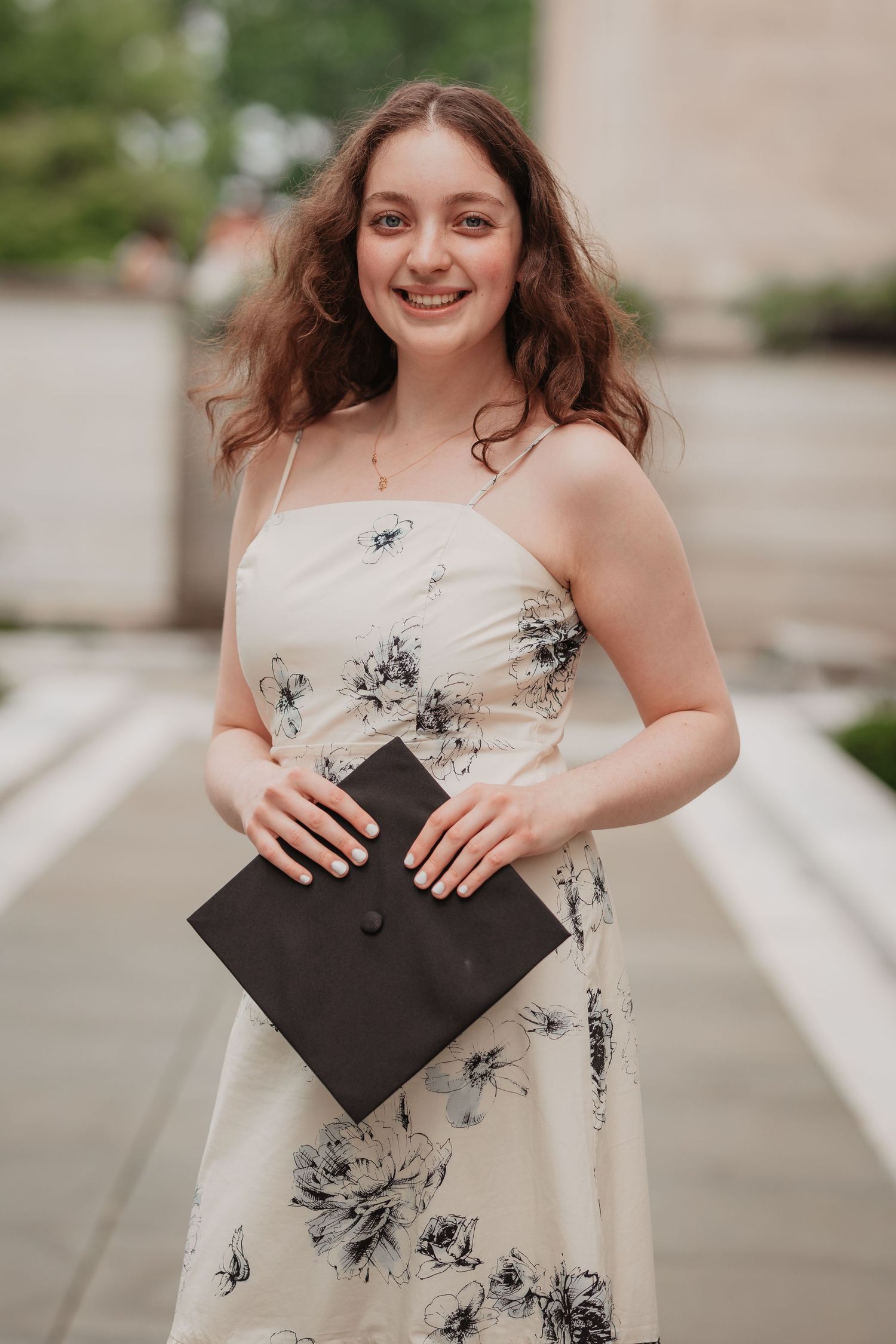 Smiling young woman in floral dress holding black graduation cap in outdoor setting.