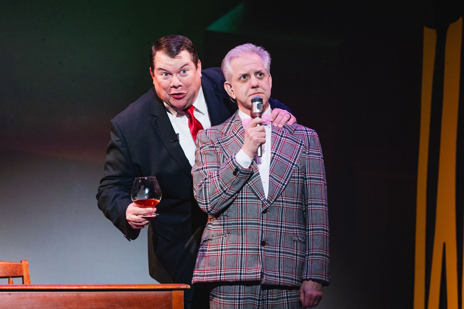 Two male performers in suits, one holding a microphone and wine glass, perform together on stage.