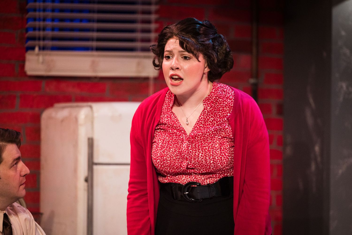 A woman in a red floral blouse and black skirt performs on a theater stage with brick backdrop.