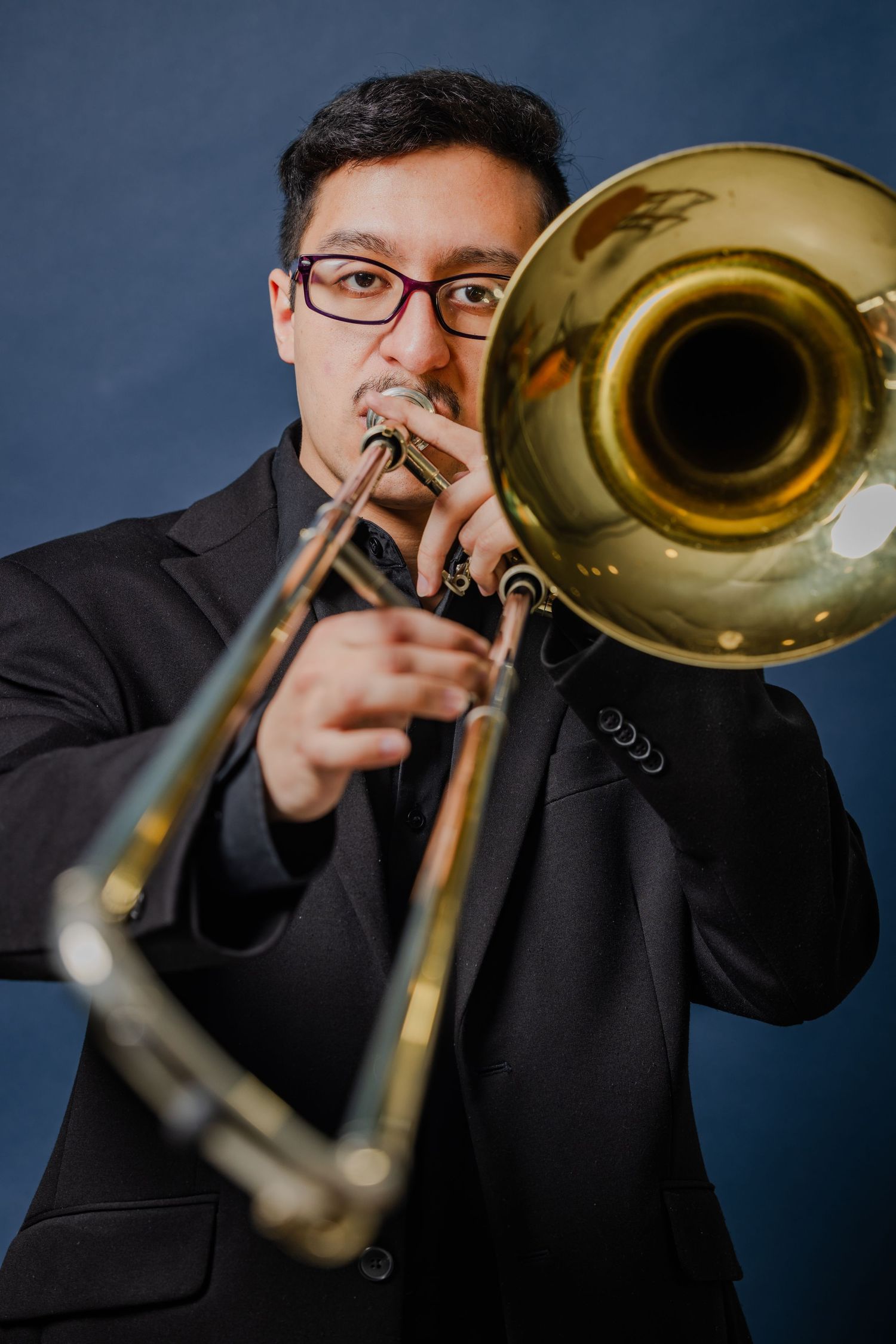 Male musician playing trombone in a black suit against a dark blue background.