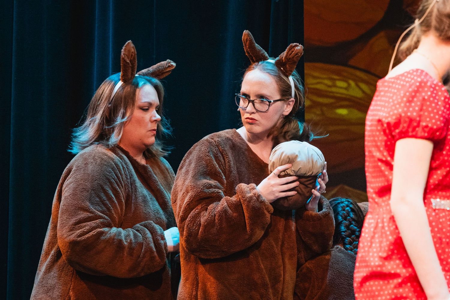 Two performers in brown bear costumes hold a prop on a theatrical stage during a school production.