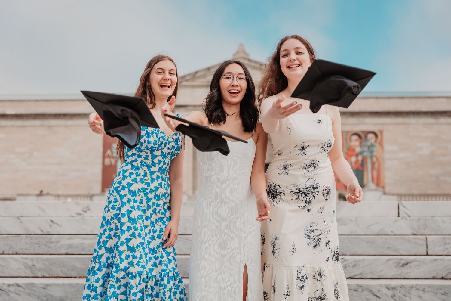 Three joyful female graduates in colorful dresses tossing mortarboard caps on museum steps.