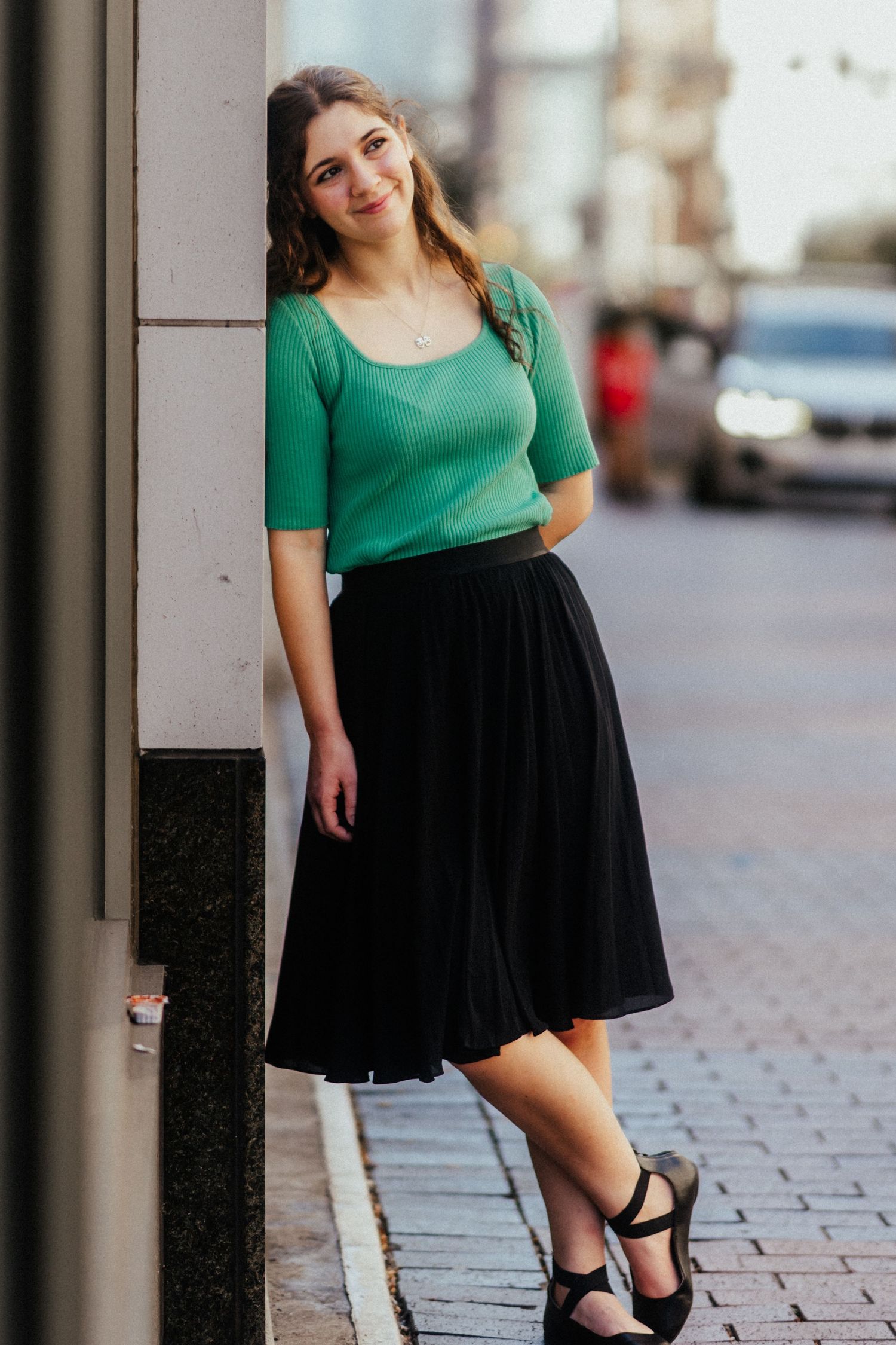 Young woman in green top and black midi skirt leans against a building wall on a city sidewalk.