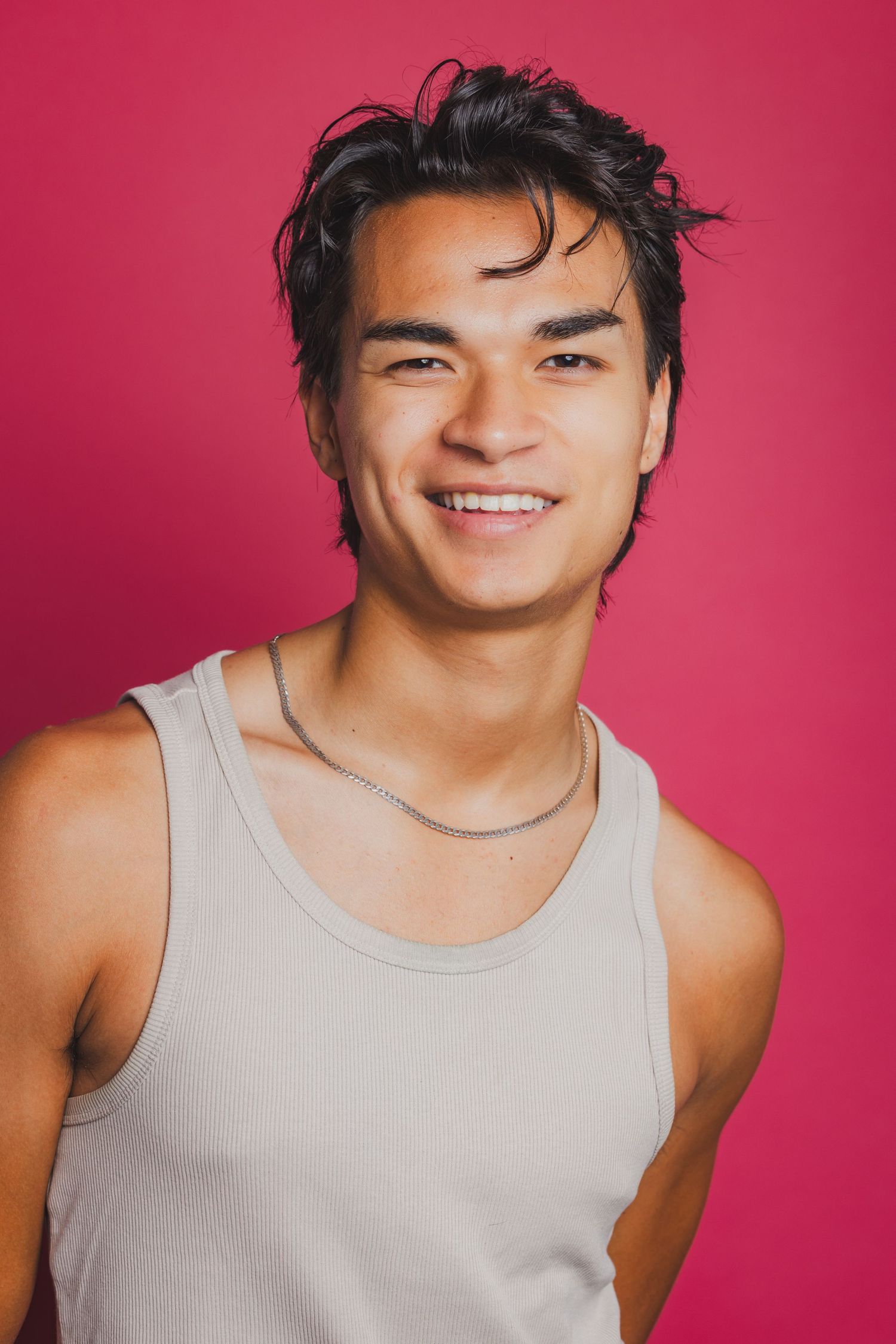 Smiling young Asian man wearing a white tank top and chain necklace posing against a pink background.