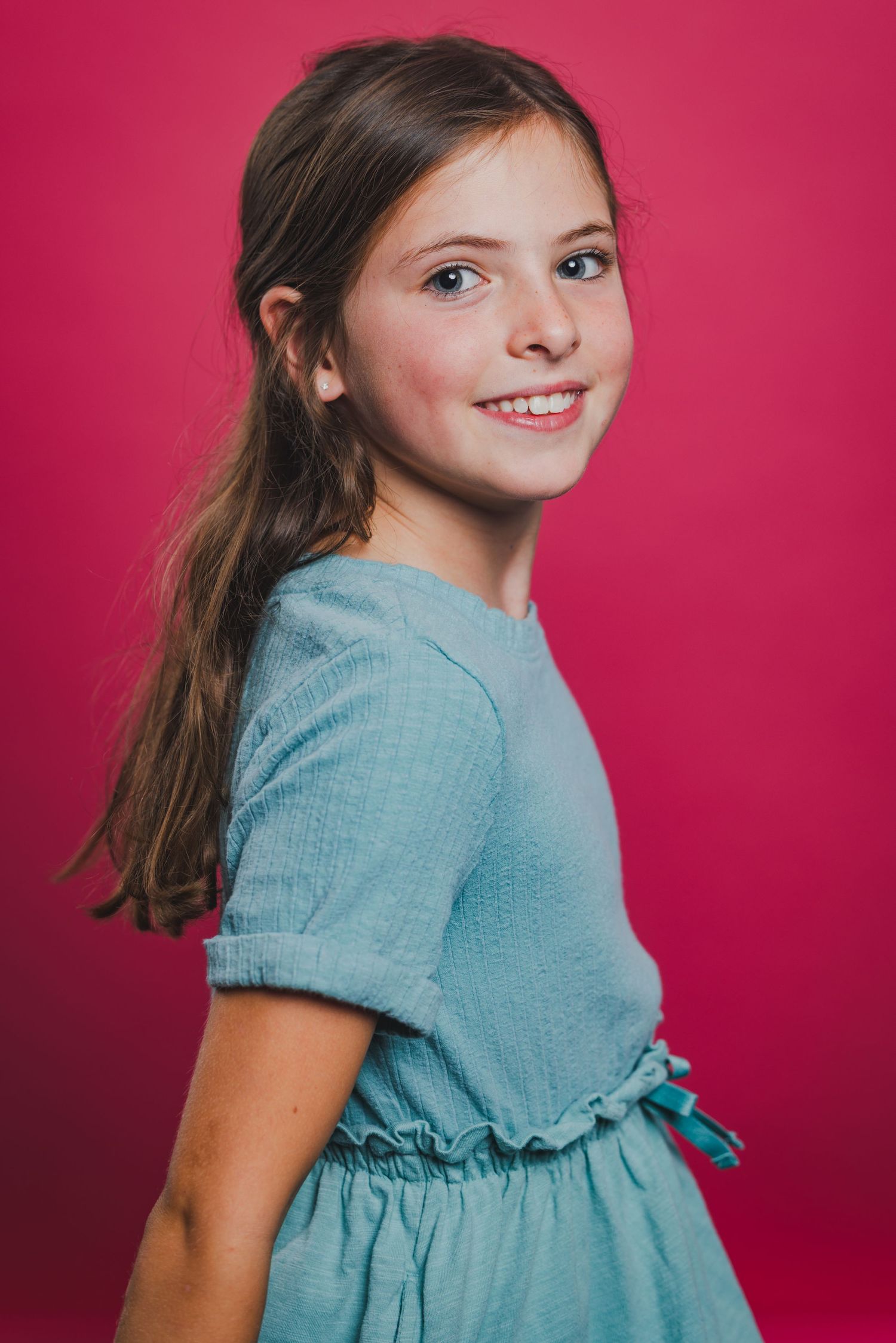 Smiling young girl with long brown hair wearing a light blue dress posed against a vibrant pink background.