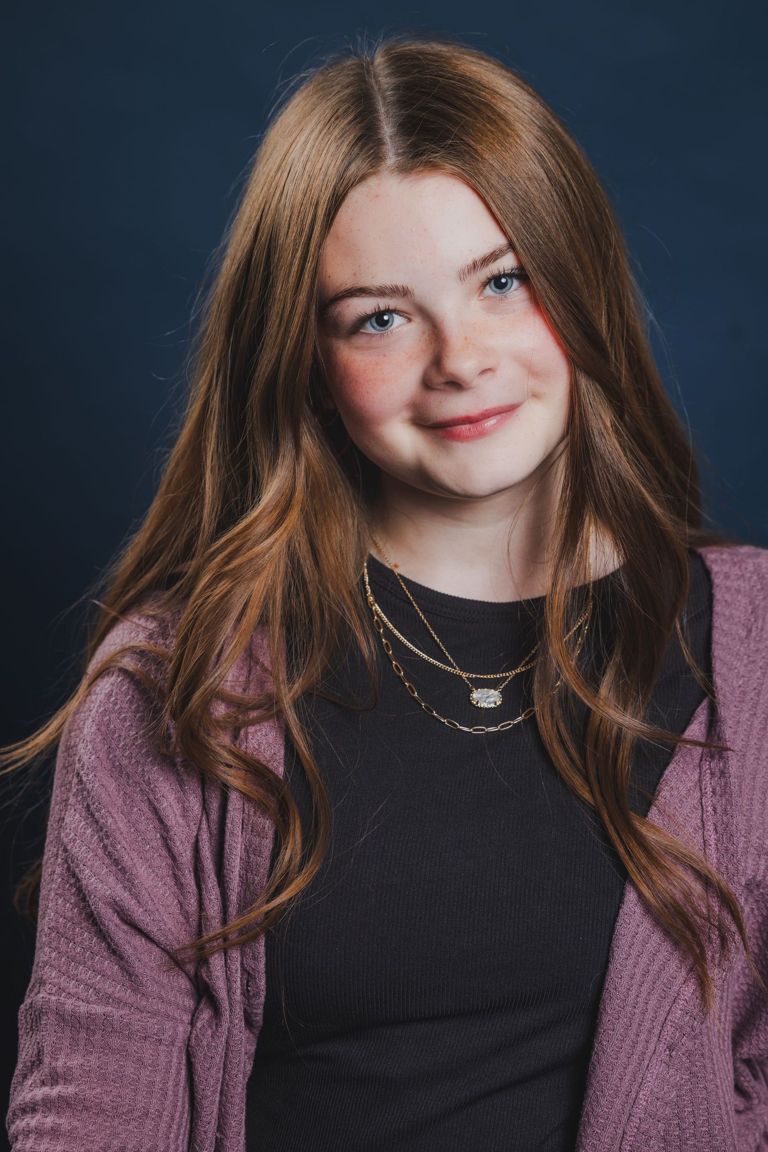 Young girl with long auburn hair smiles warmly wearing a pink cardigan and layered necklaces.