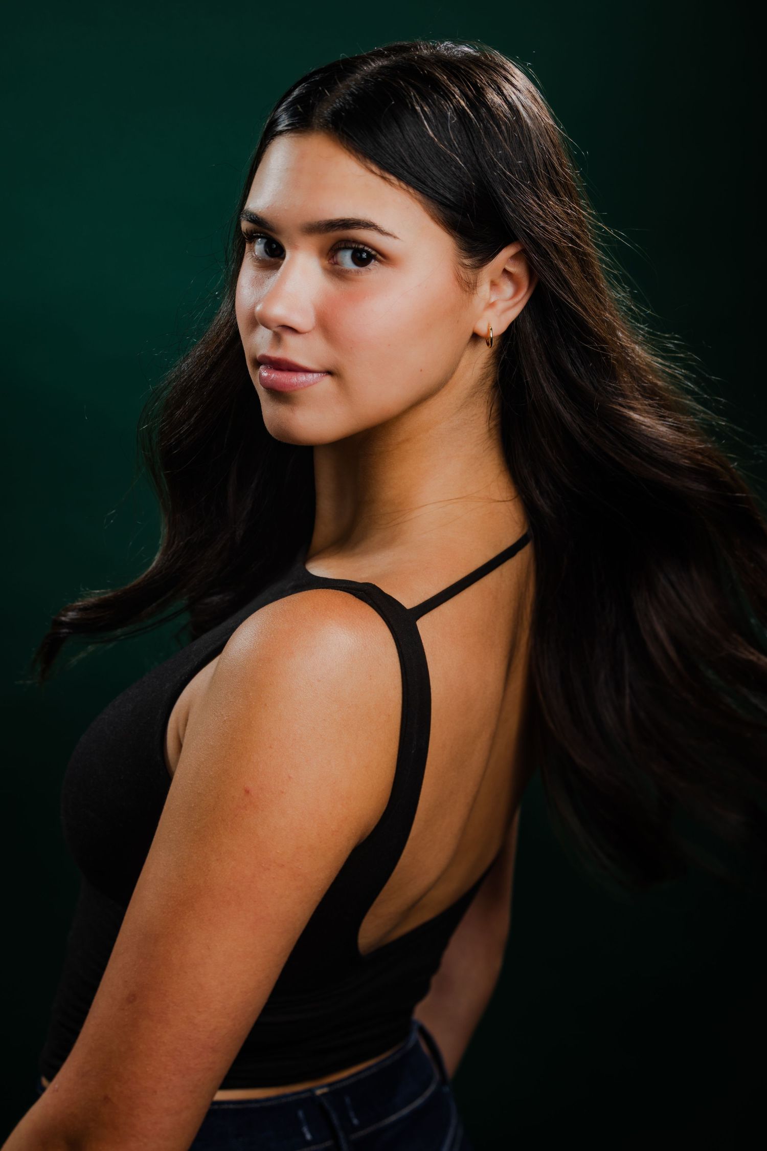 Young woman with long dark hair in a black open-back top posing over her shoulder on dark green background.