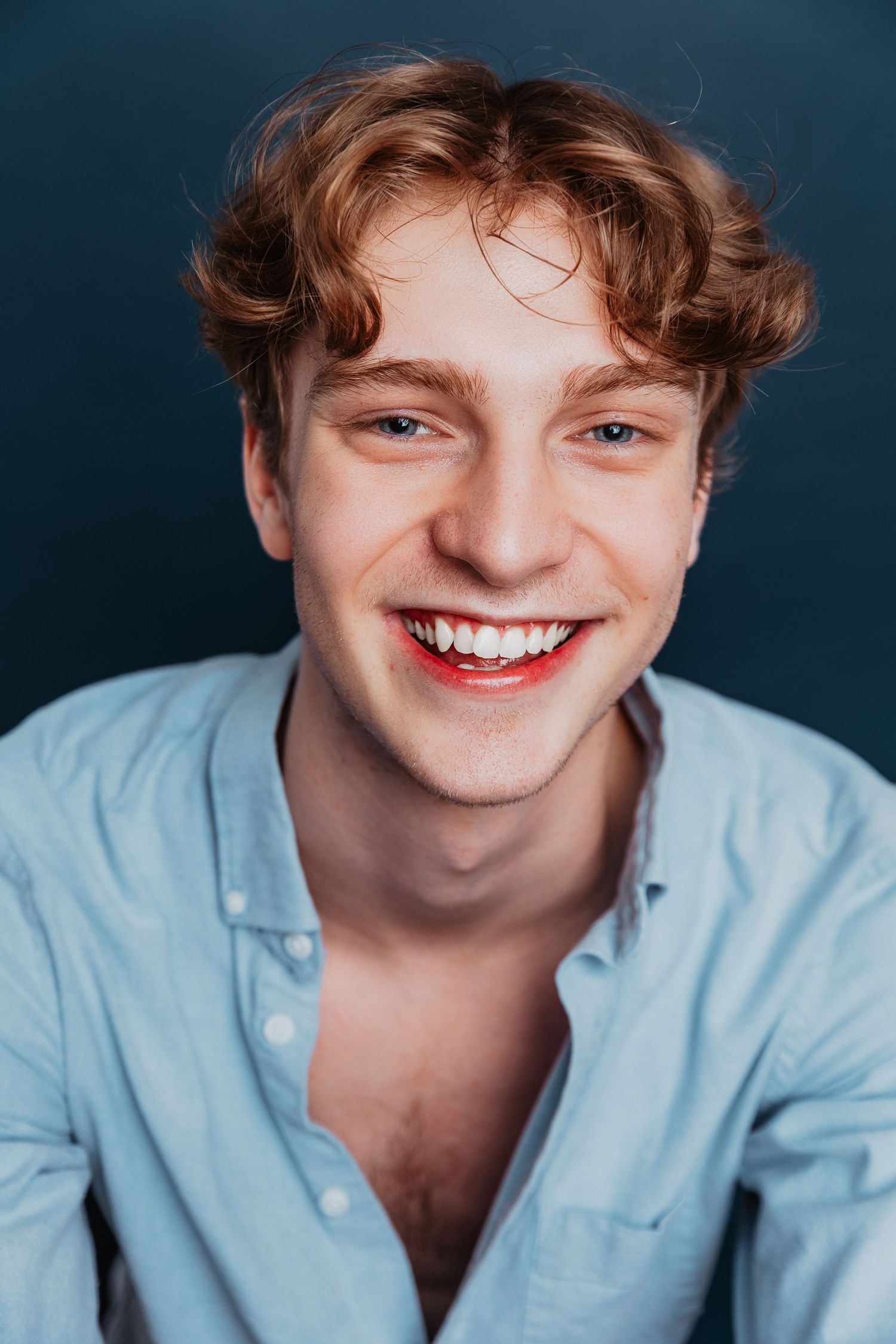 Smiling young man with curly auburn hair wearing light blue shirt against dark blue background.