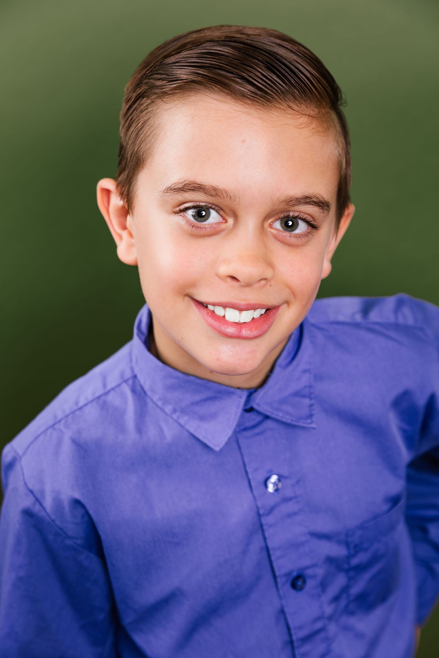 Young boy in blue button-up shirt smiling against dark green background.