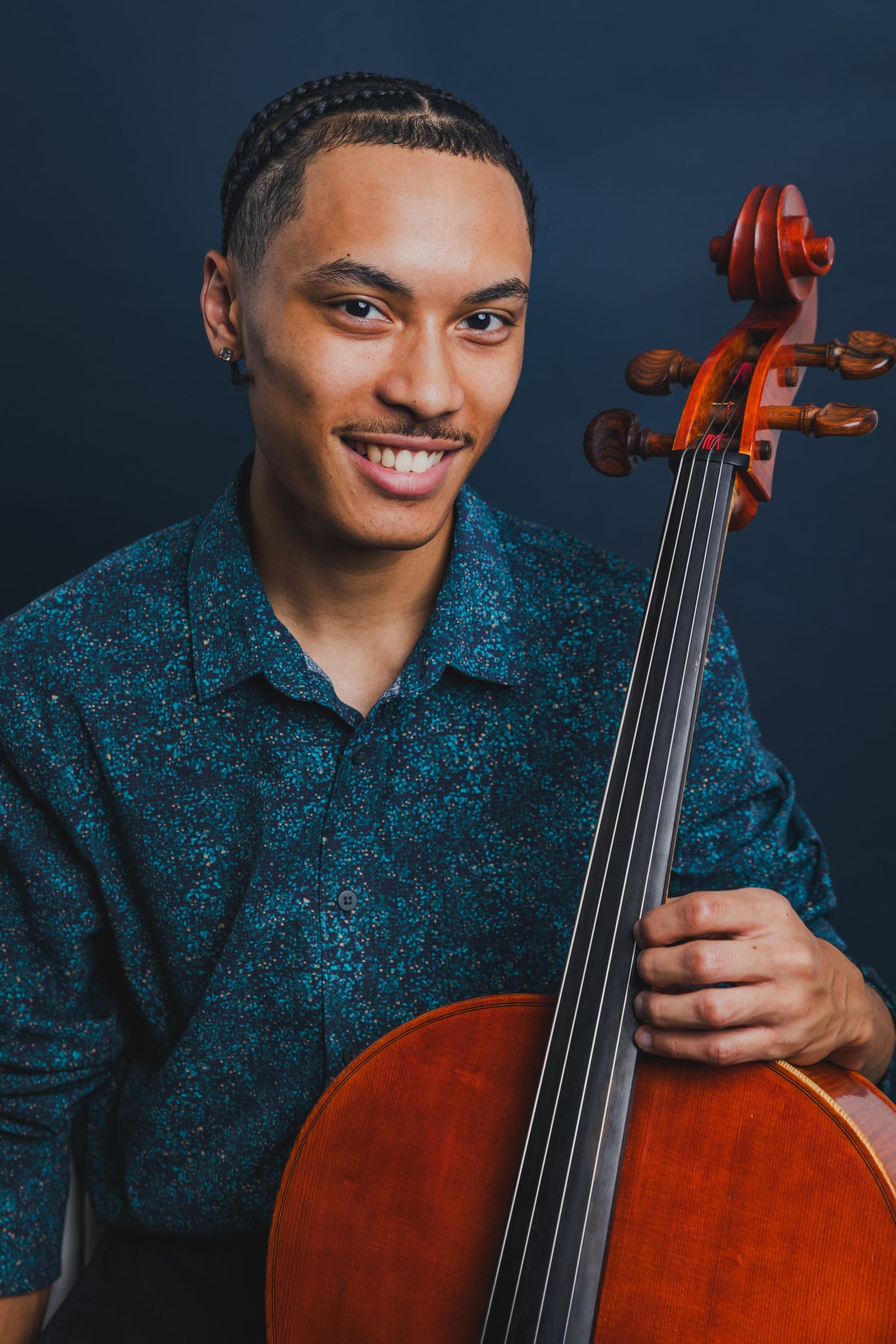 Smiling young male cellist in a teal patterned shirt holding his cello against a dark navy studio background.