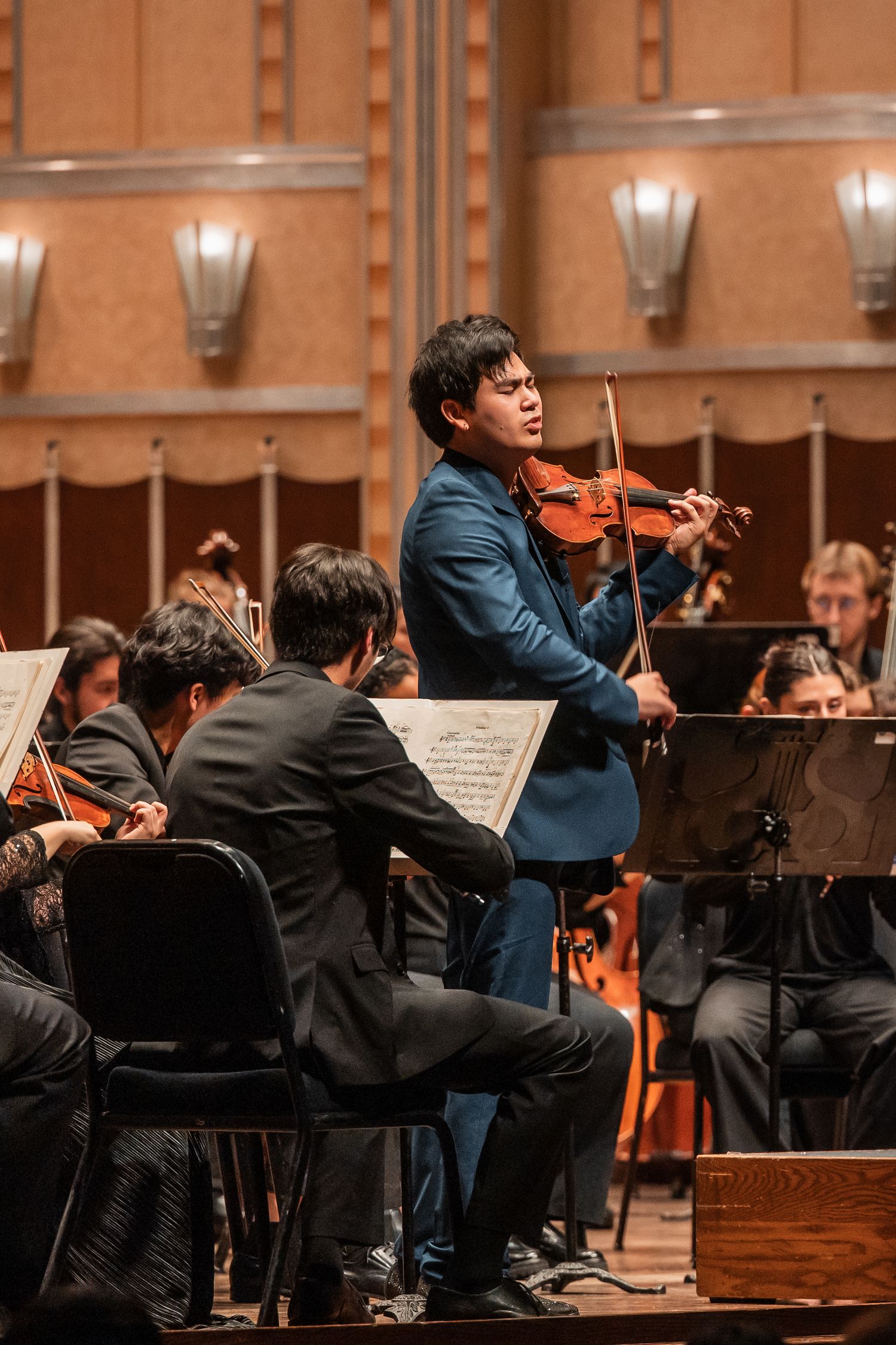 A violinist in a blue suit performs as a soloist with a full orchestra in a grand concert hall.