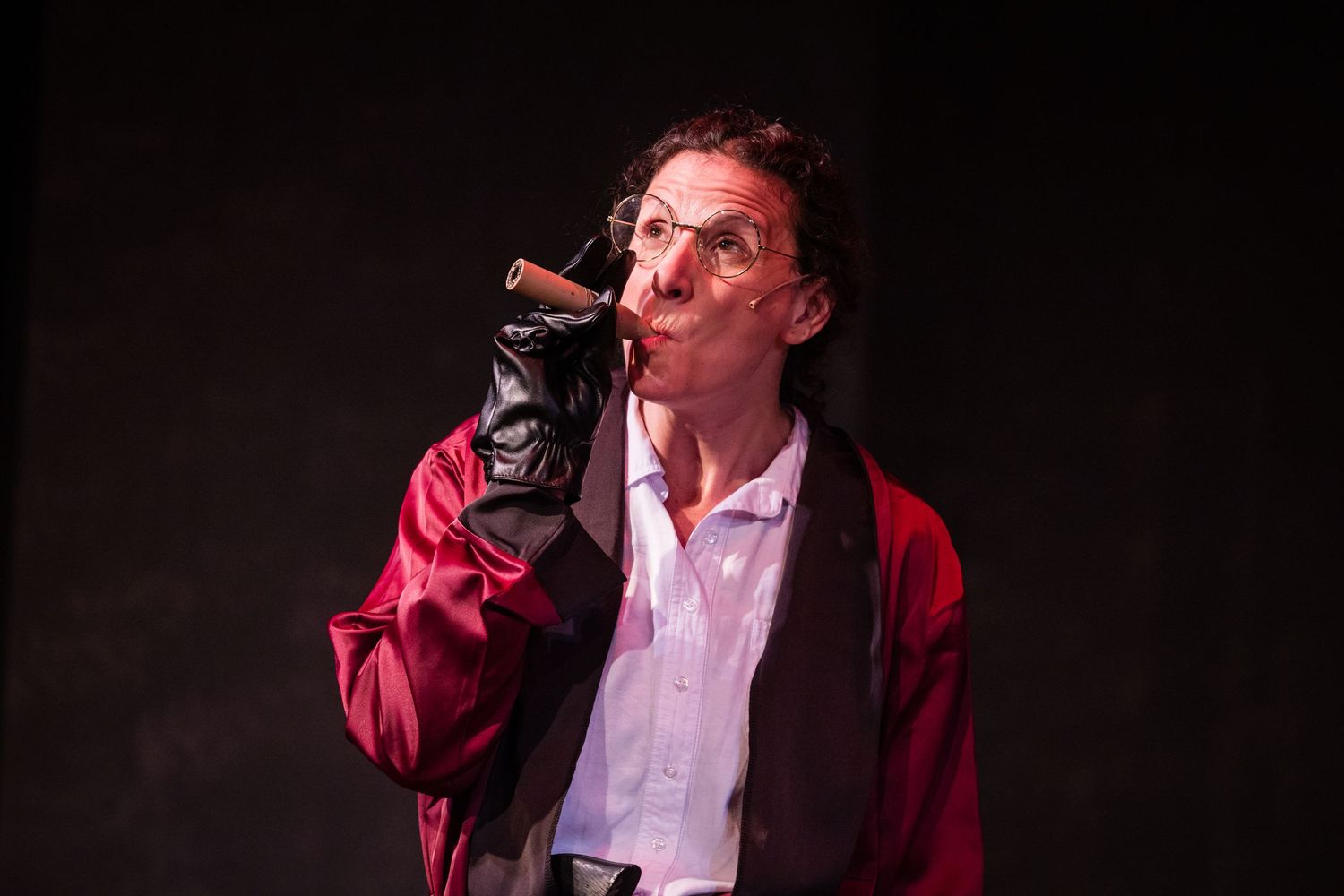 Theatrical performer in a red jacket dramatically smoking a cigar against a dark studio background.