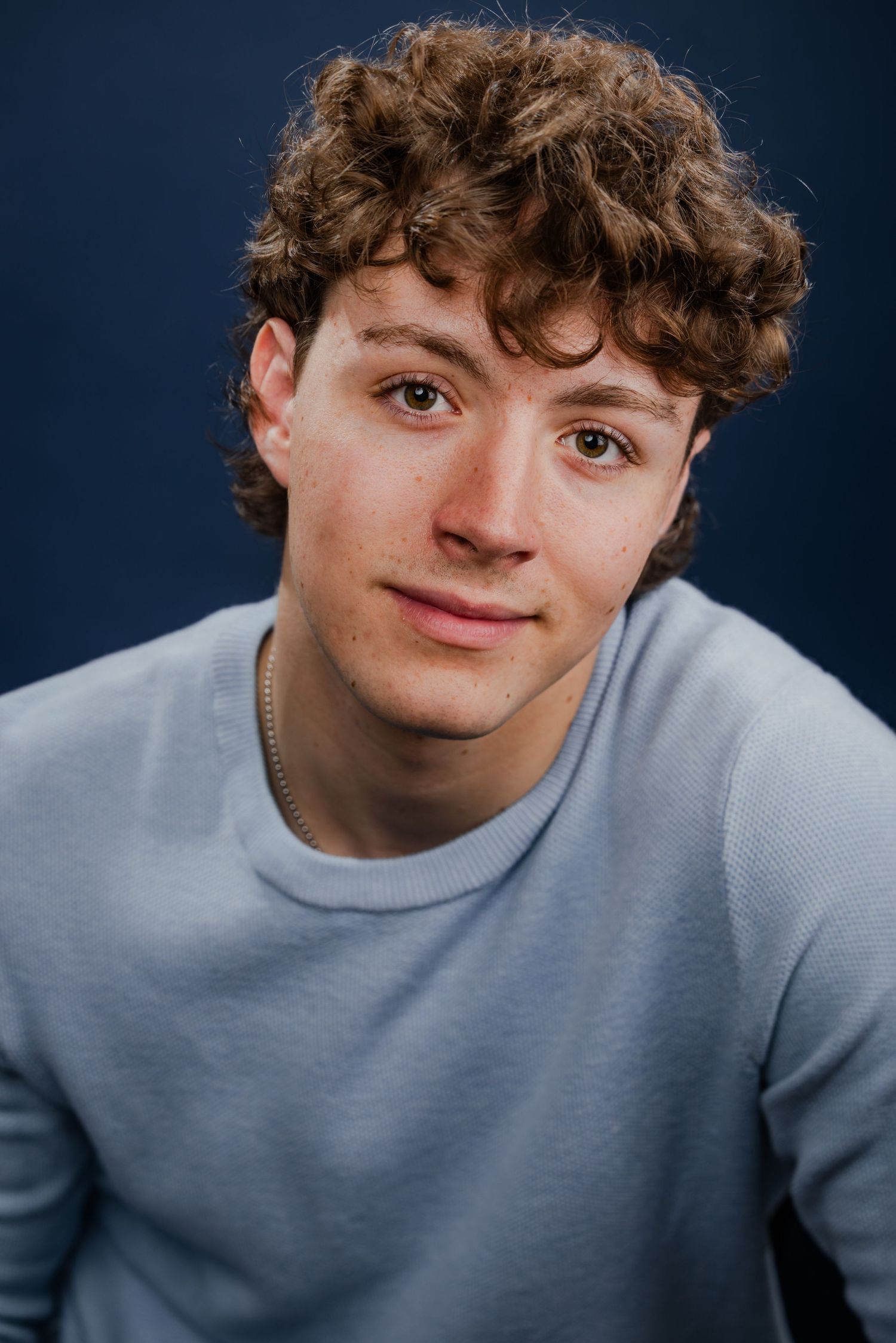 Young man with curly hair in a light blue sweatshirt posing against a dark blue background.
