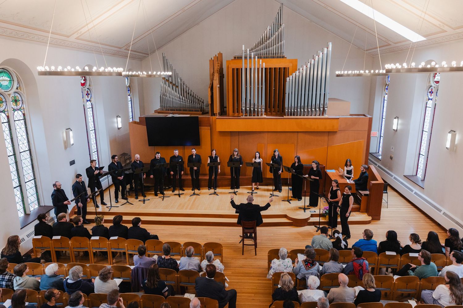 Choral ensemble performs in a bright white chapel with pipe organ while conductor leads from center aisle.