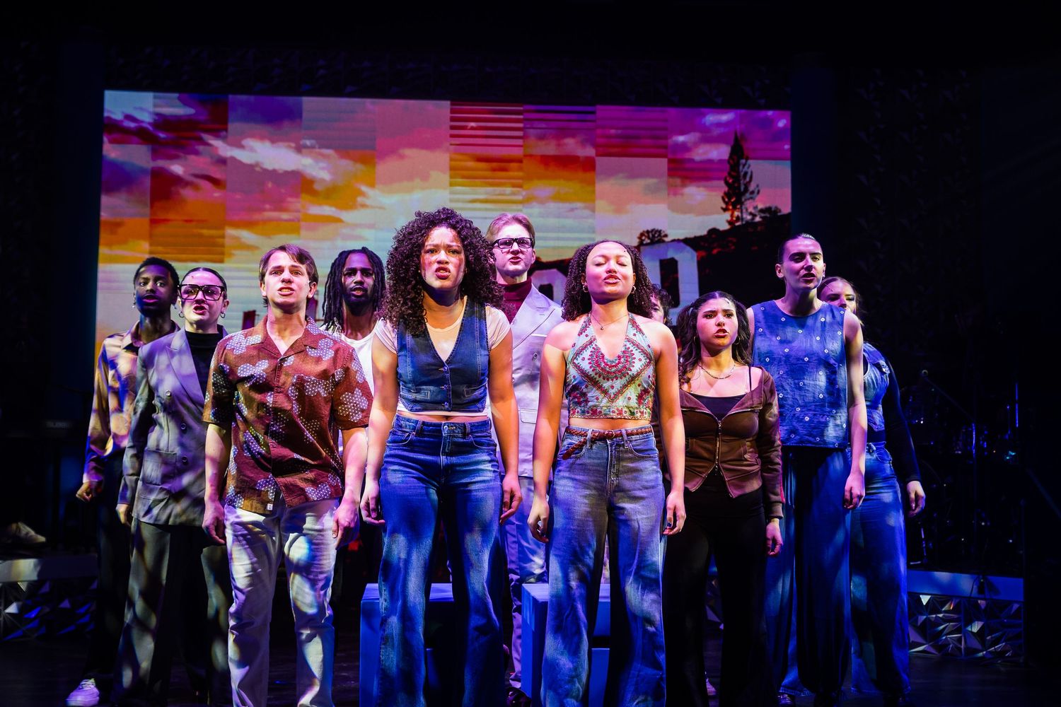 Diverse theater cast stands together dramatically on stage with colorful sunset backdrop projection.