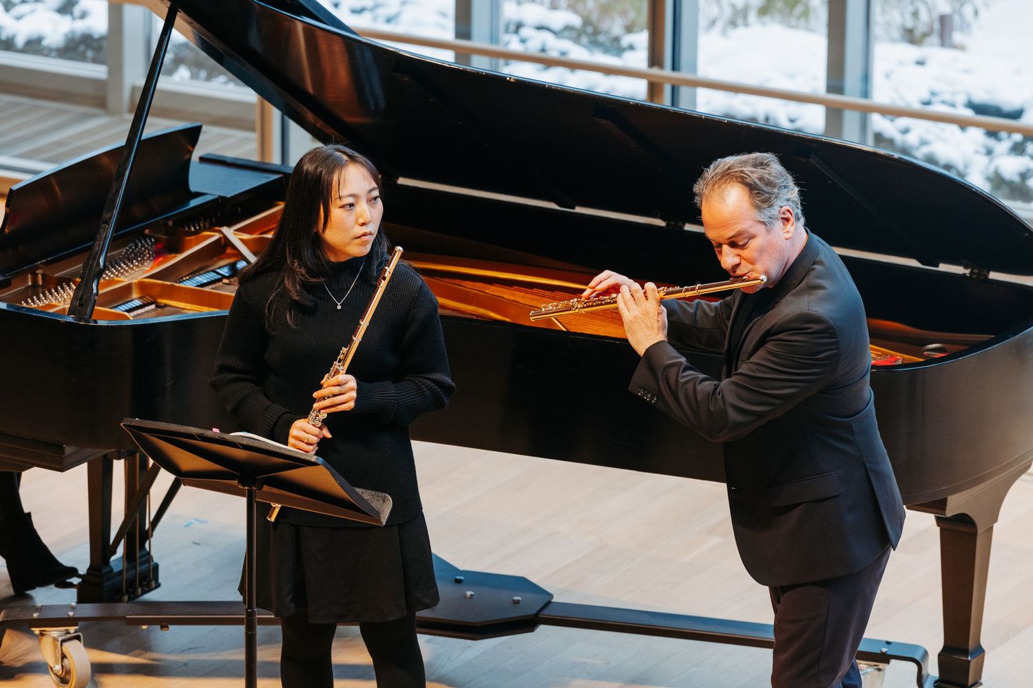 A flutist and pianist rehearse together in a bright modern performance space with a grand piano.