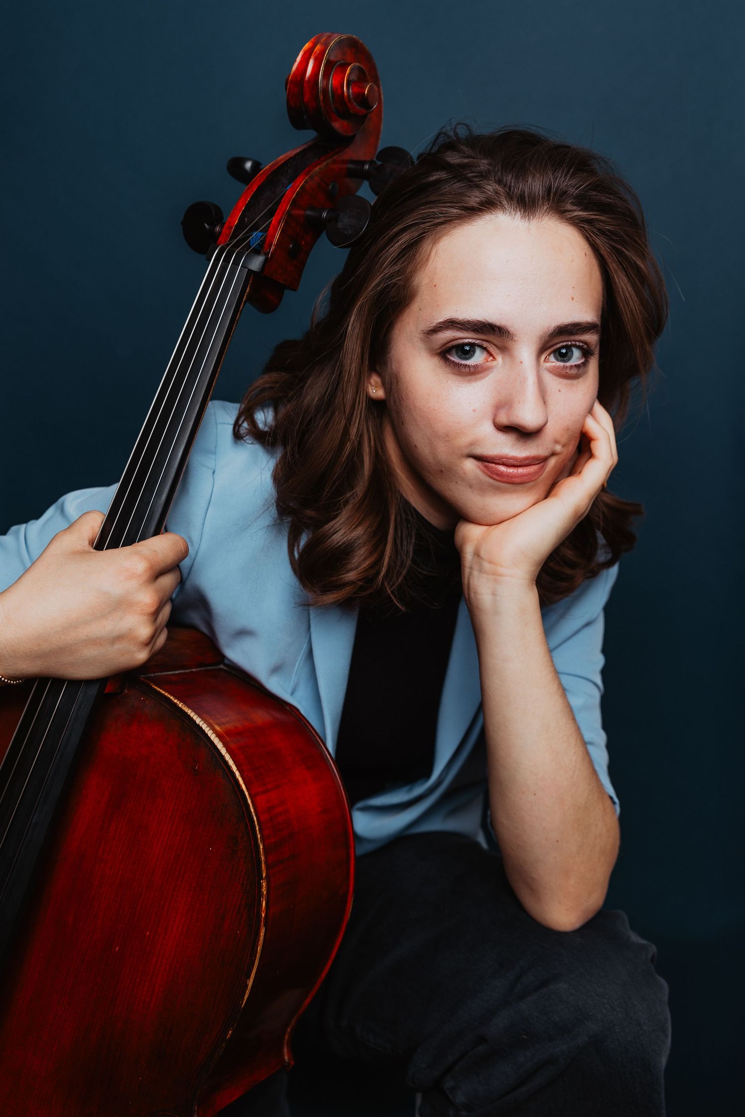 Thoughtful young musician resting chin on hand while holding a cello, seated against a dark blue background.
