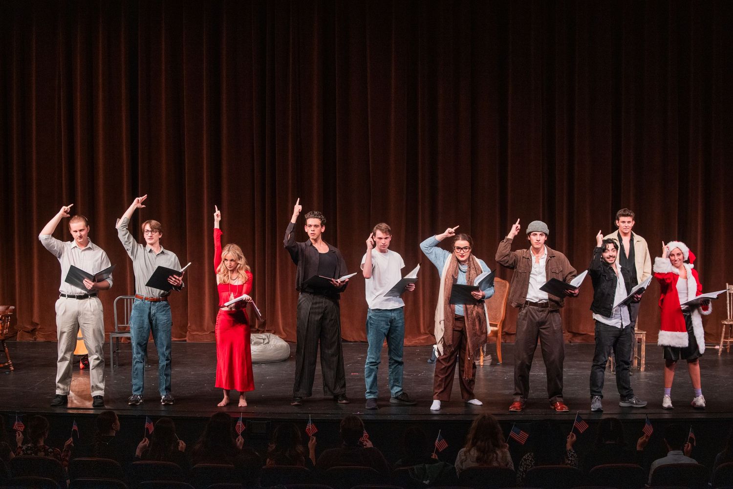Theater cast of eight performers taking a bow on stage holding scripts after a performance.