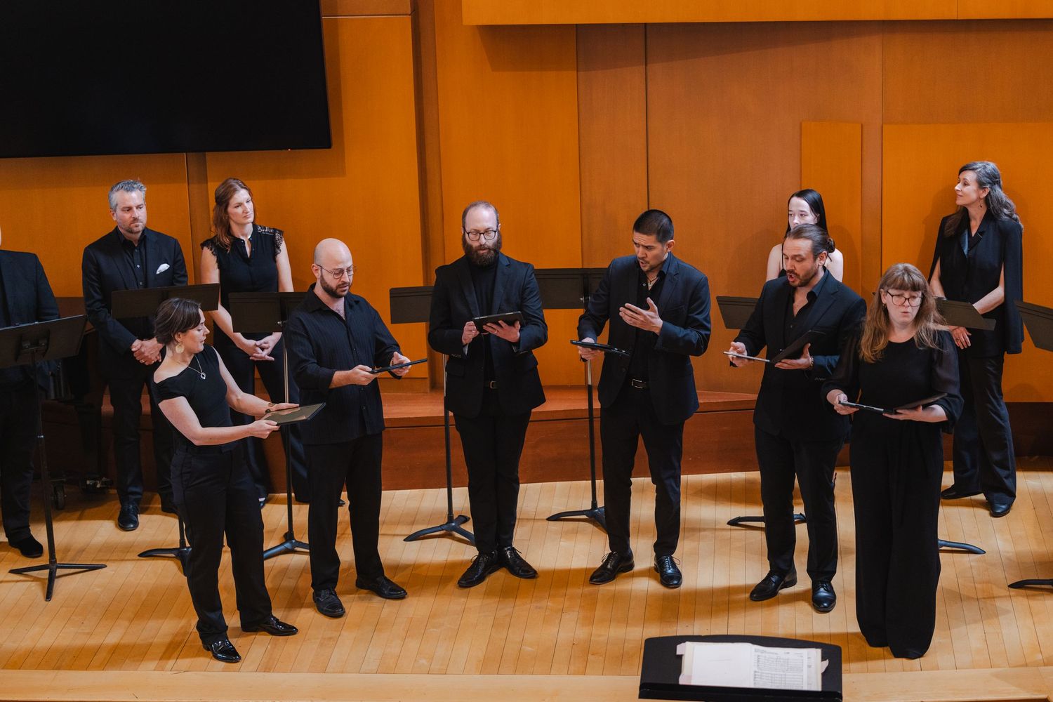 Vocal ensemble of ten singers dressed in black performing on a warm wood-paneled concert stage.