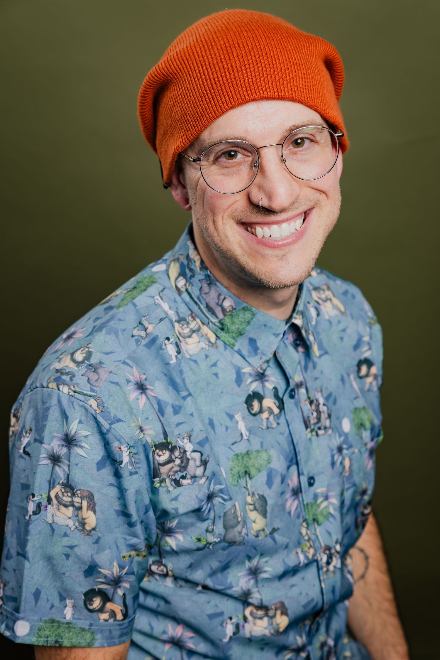 Smiling man wearing orange beanie, glasses, and tropical print shirt, olive background portrait.