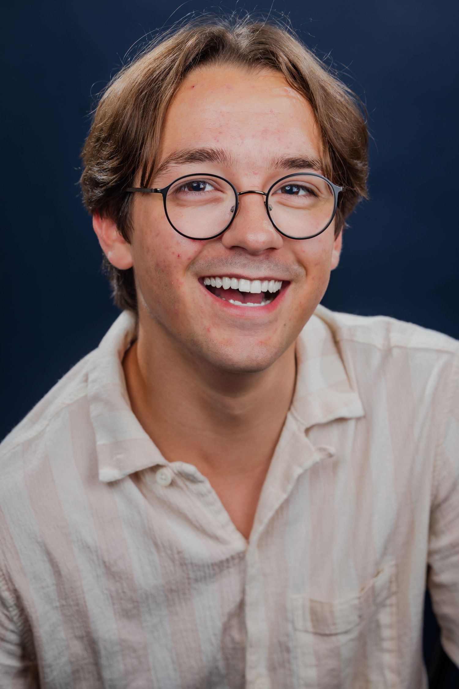 Smiling young man with glasses wearing a beige striped shirt against a dark blue background.