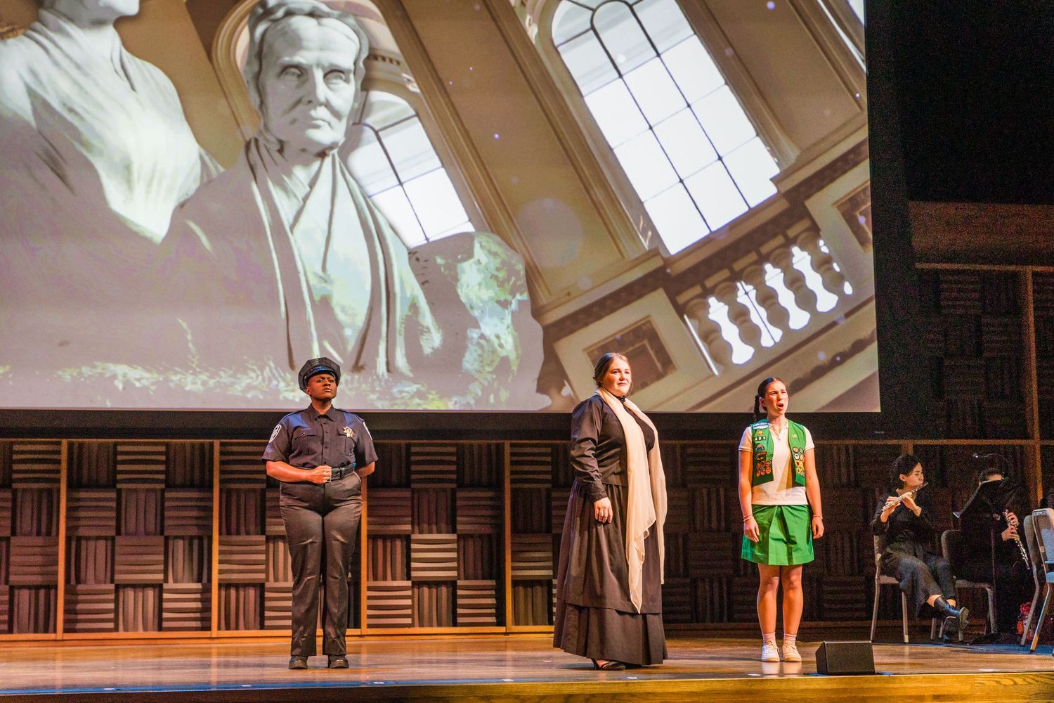 Three performers stand on stage beneath a projected architectural image during a theatrical performance.