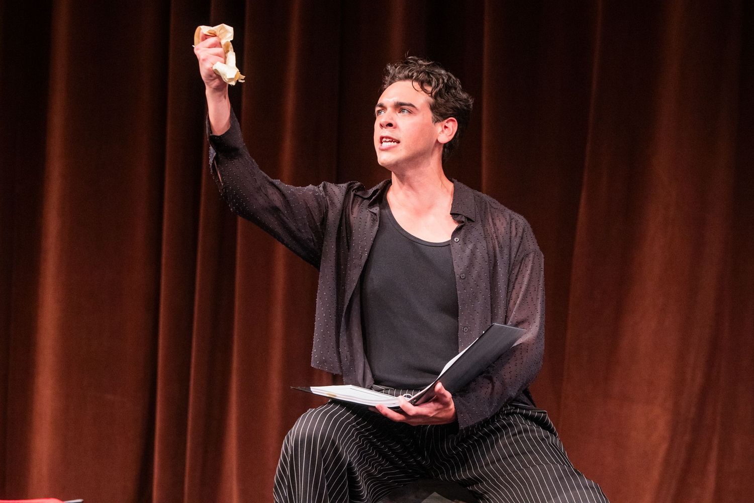 A male performer holds script and raises hand dramatically on stage with red curtain backdrop.
