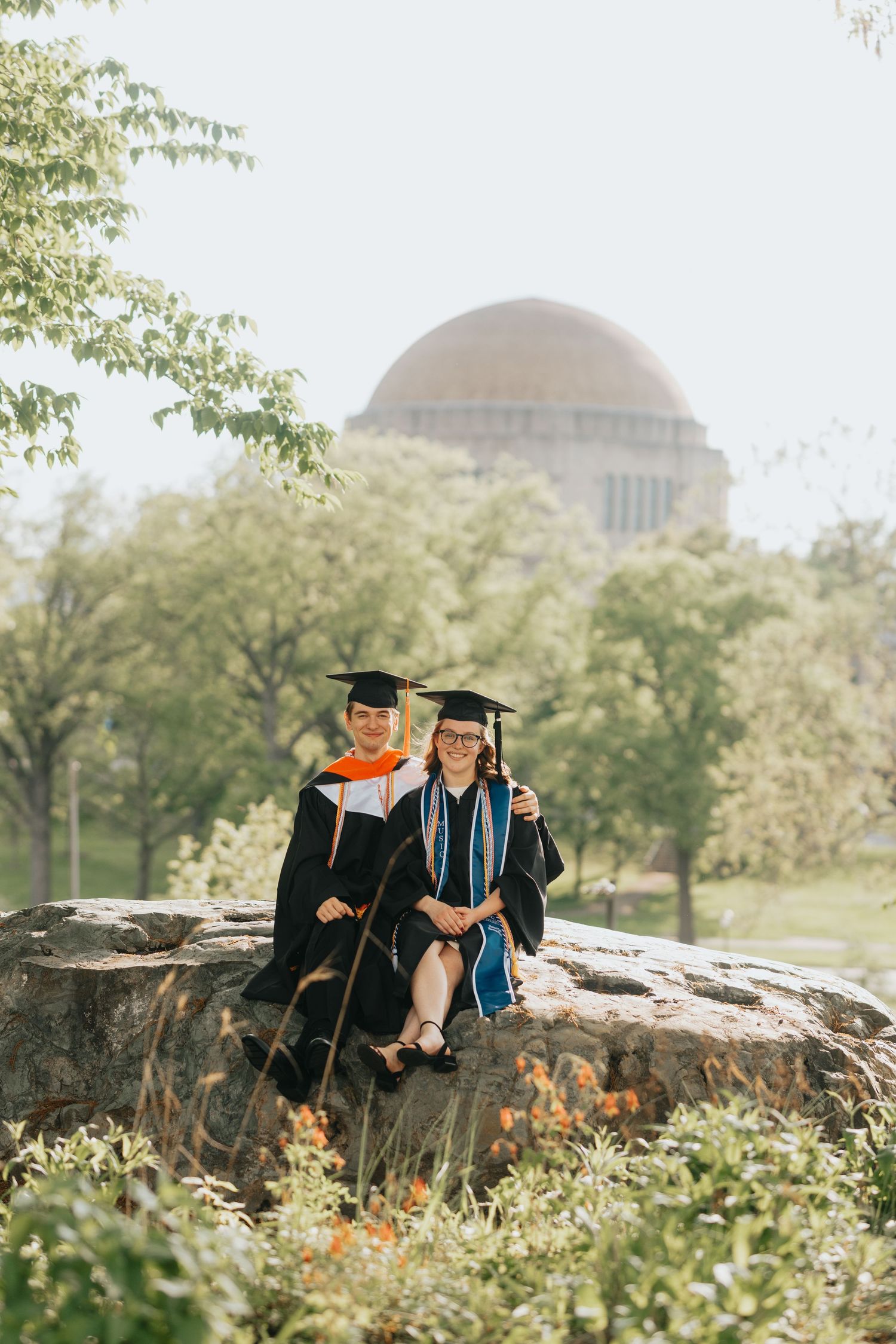 Two female graduates in caps and gowns sitting on rocks with domed building behind them.