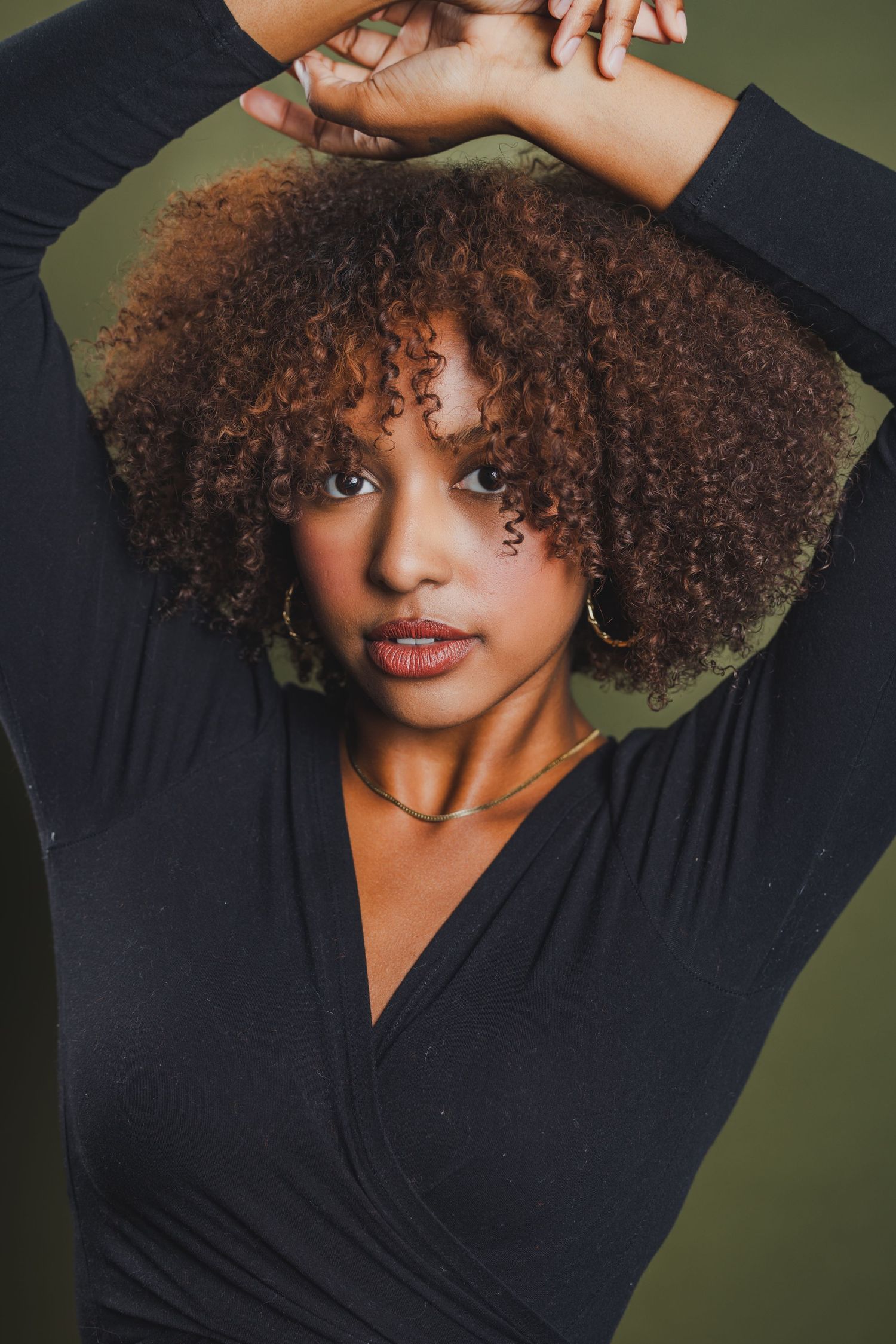 Woman with curly brown hair in black wrap top poses with arms raised against olive green background.