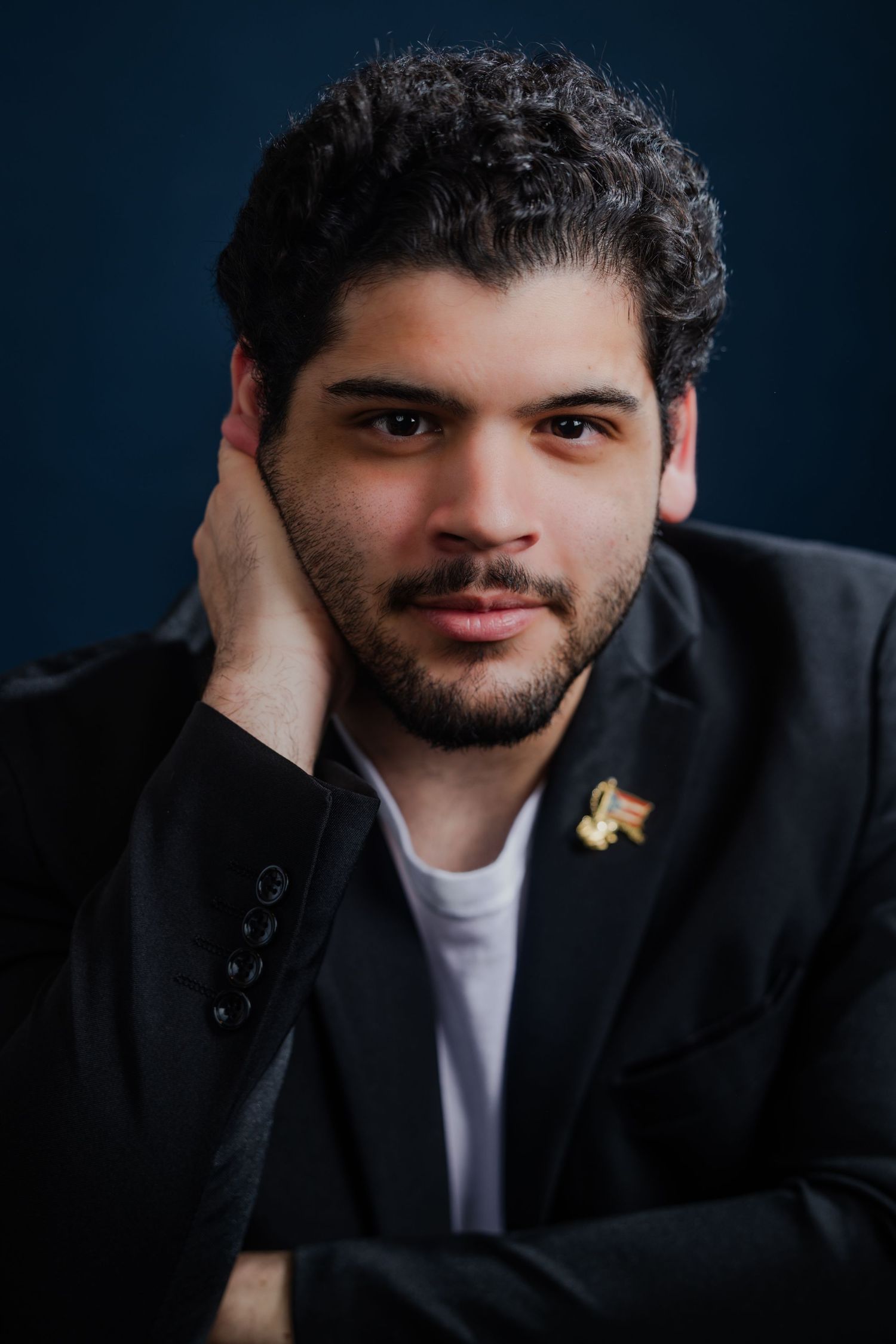 Young man with curly dark hair in a black blazer posing against a dark blue background.