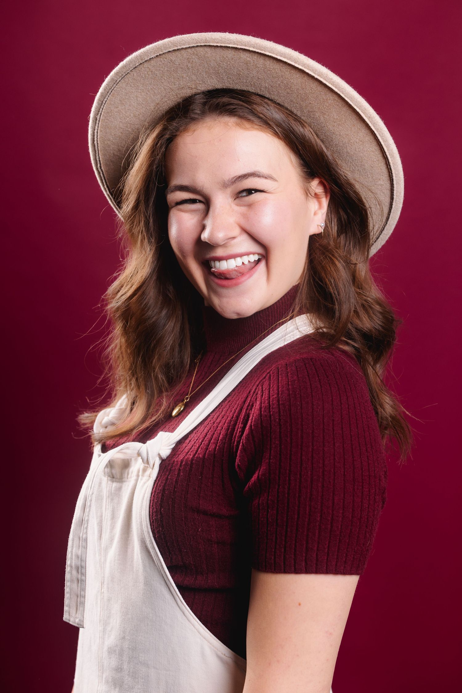 Laughing young woman in a hat and overalls posing against a deep red background.