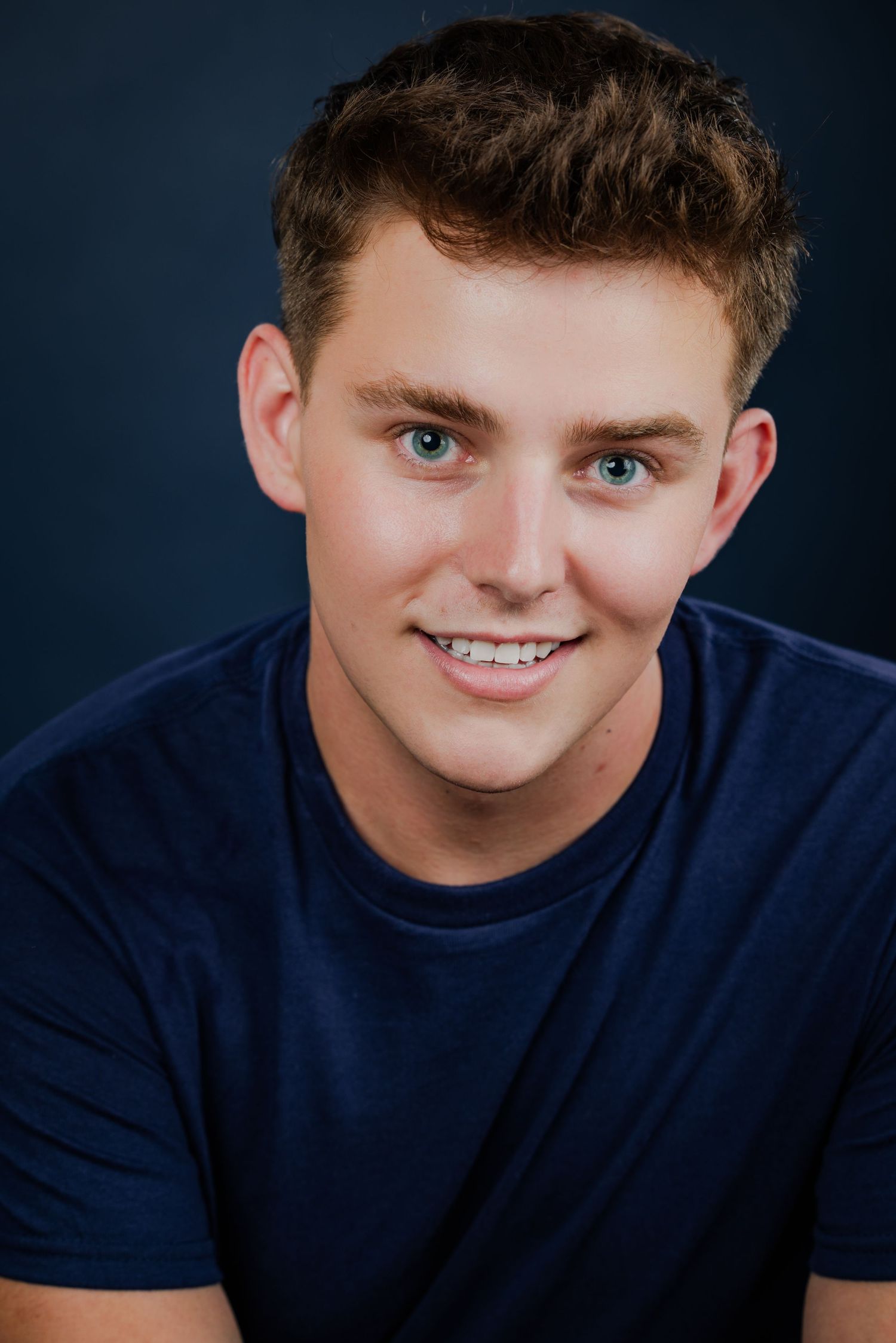 Smiling young man with blue eyes and braces wearing a navy t-shirt against a dark blue background.