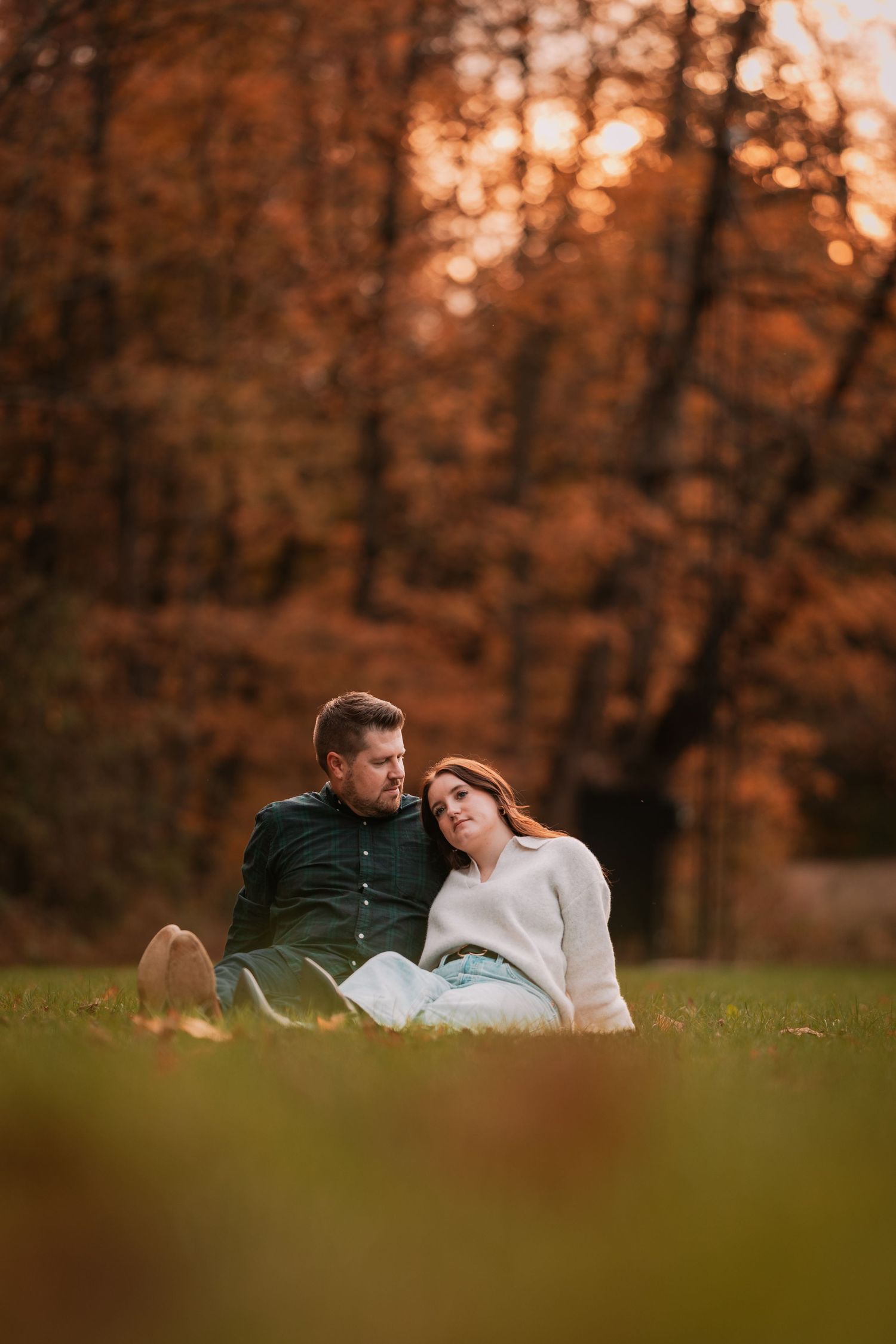 A romantic couple sits together on grass, leaning close against a backdrop of autumn trees at sunset.
