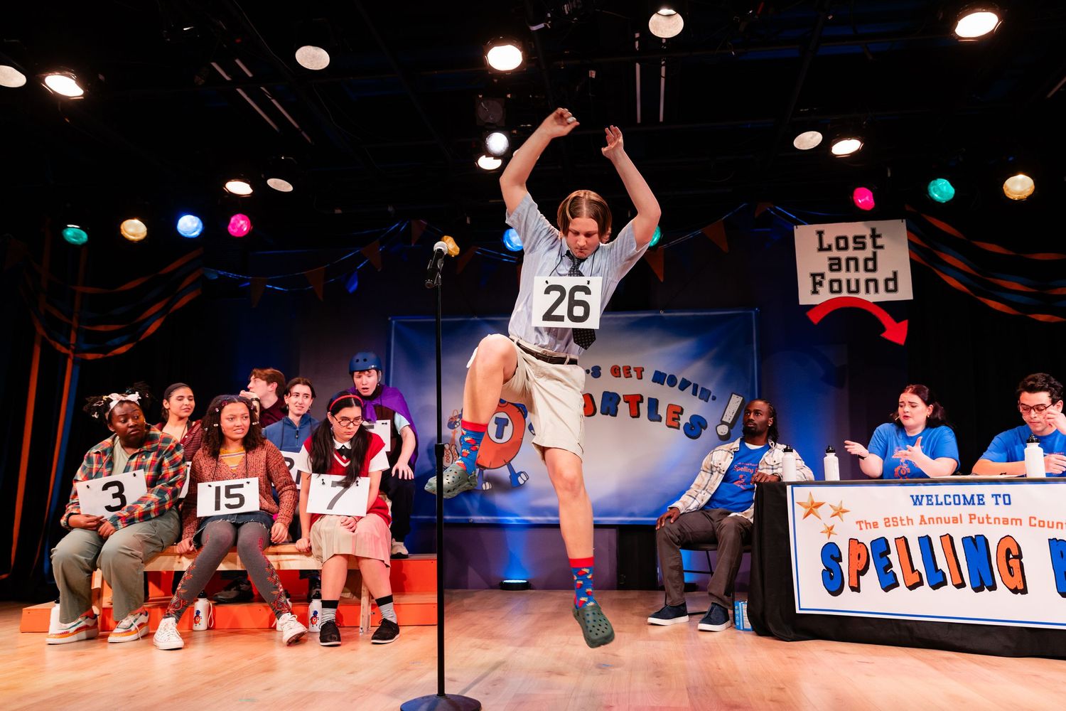 Excited performer jumping at a spelling bee competition on stage with colorful lights and audience in background.
