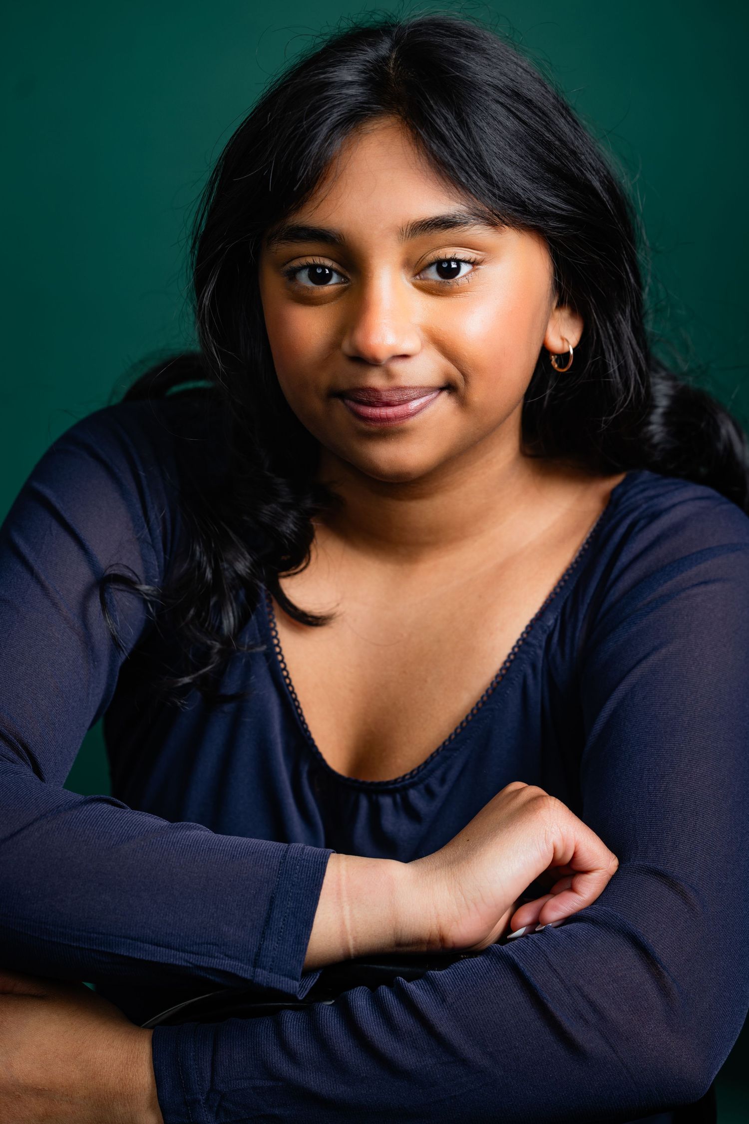 Young South Asian woman in a navy blue top with arms crossed, smiling against a dark green background.