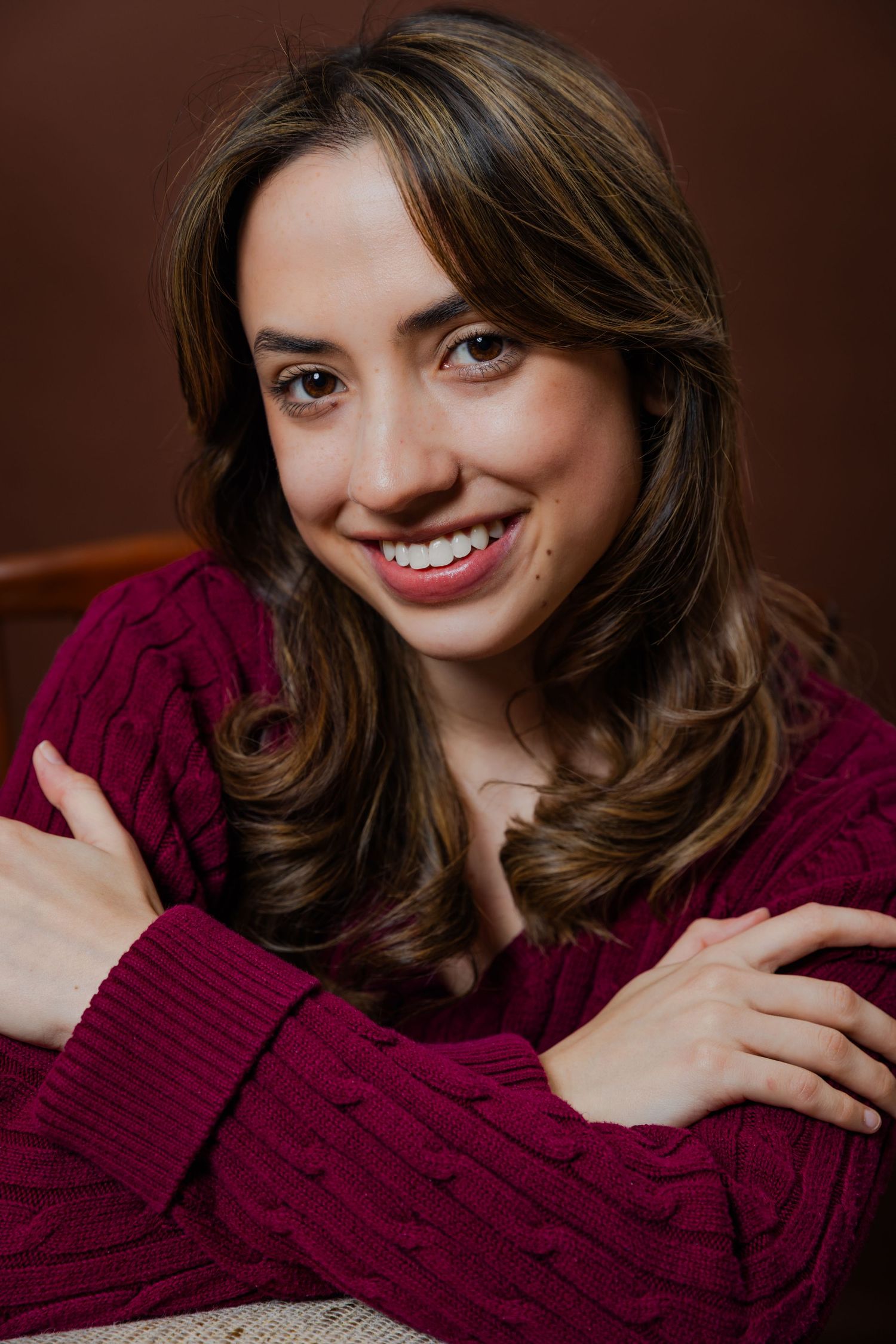 A young woman with brown hair wearing a burgundy cable-knit sweater smiles against a brown background.