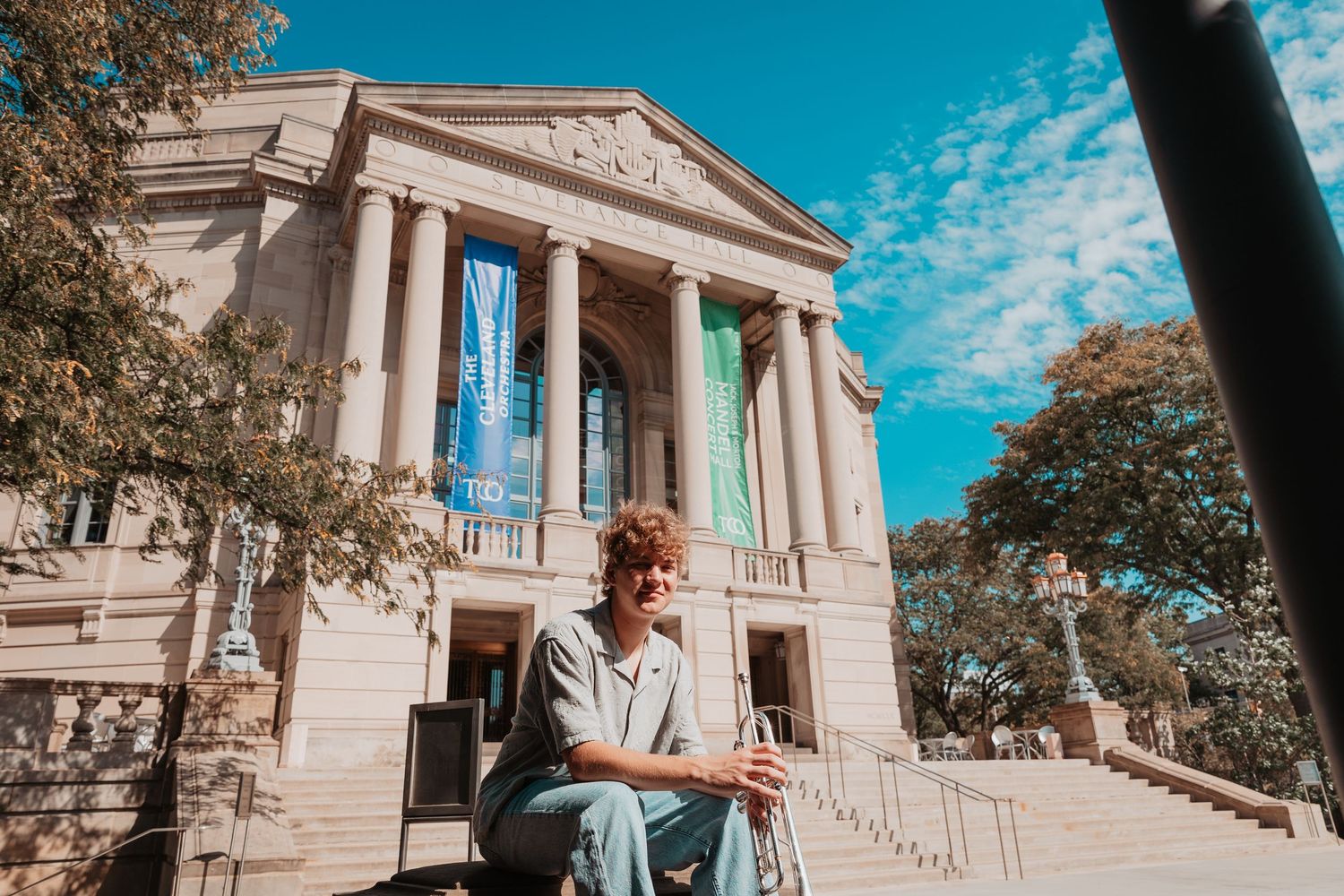 Young person sitting on steps of a neoclassical building with blue and green banners under a blue sky.