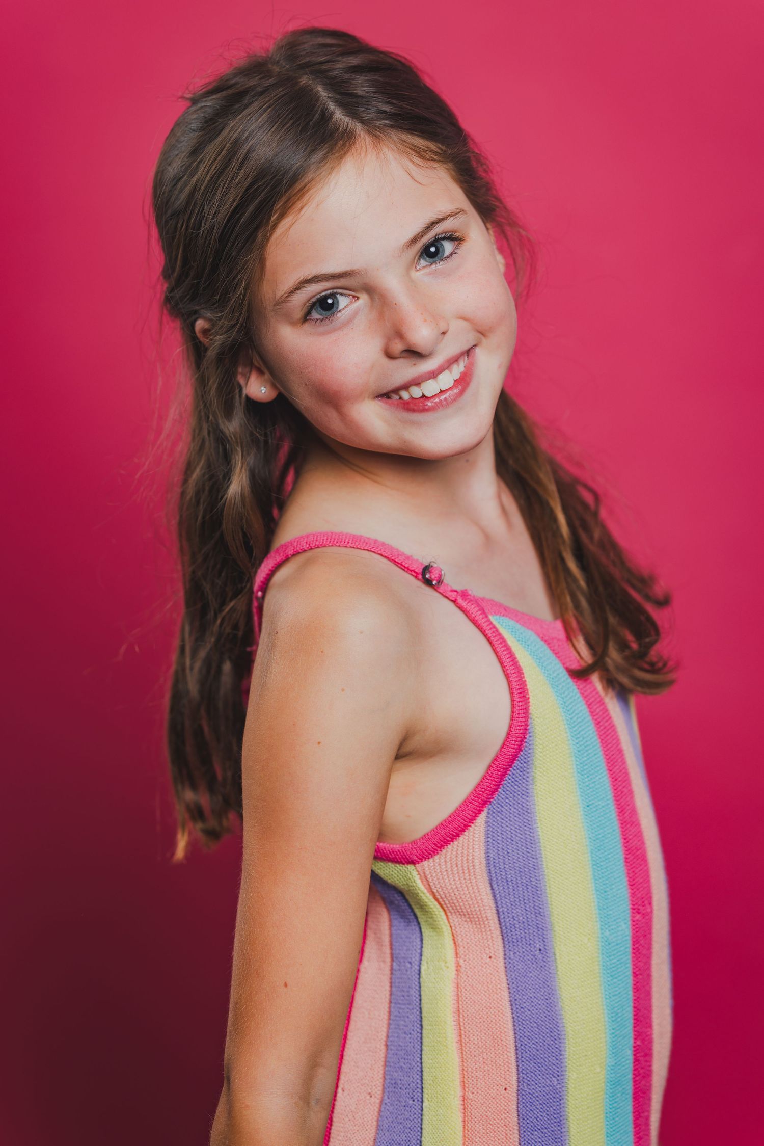 Young smiling girl with brown hair wearing a colorful striped sundress, posing against a bright pink background.