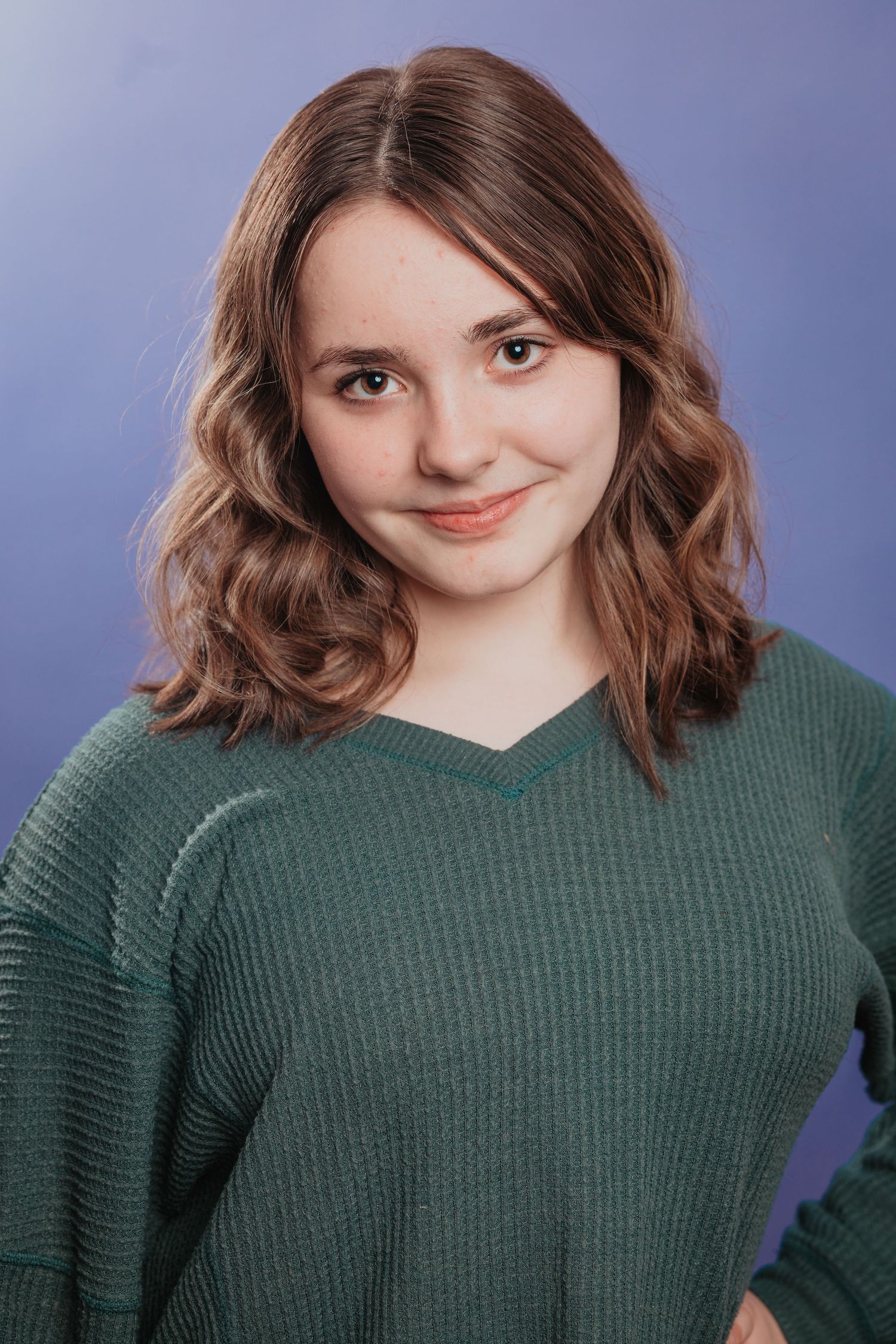 Teenage girl with wavy brown hair smiling softly in a green sweater against a purple background.
