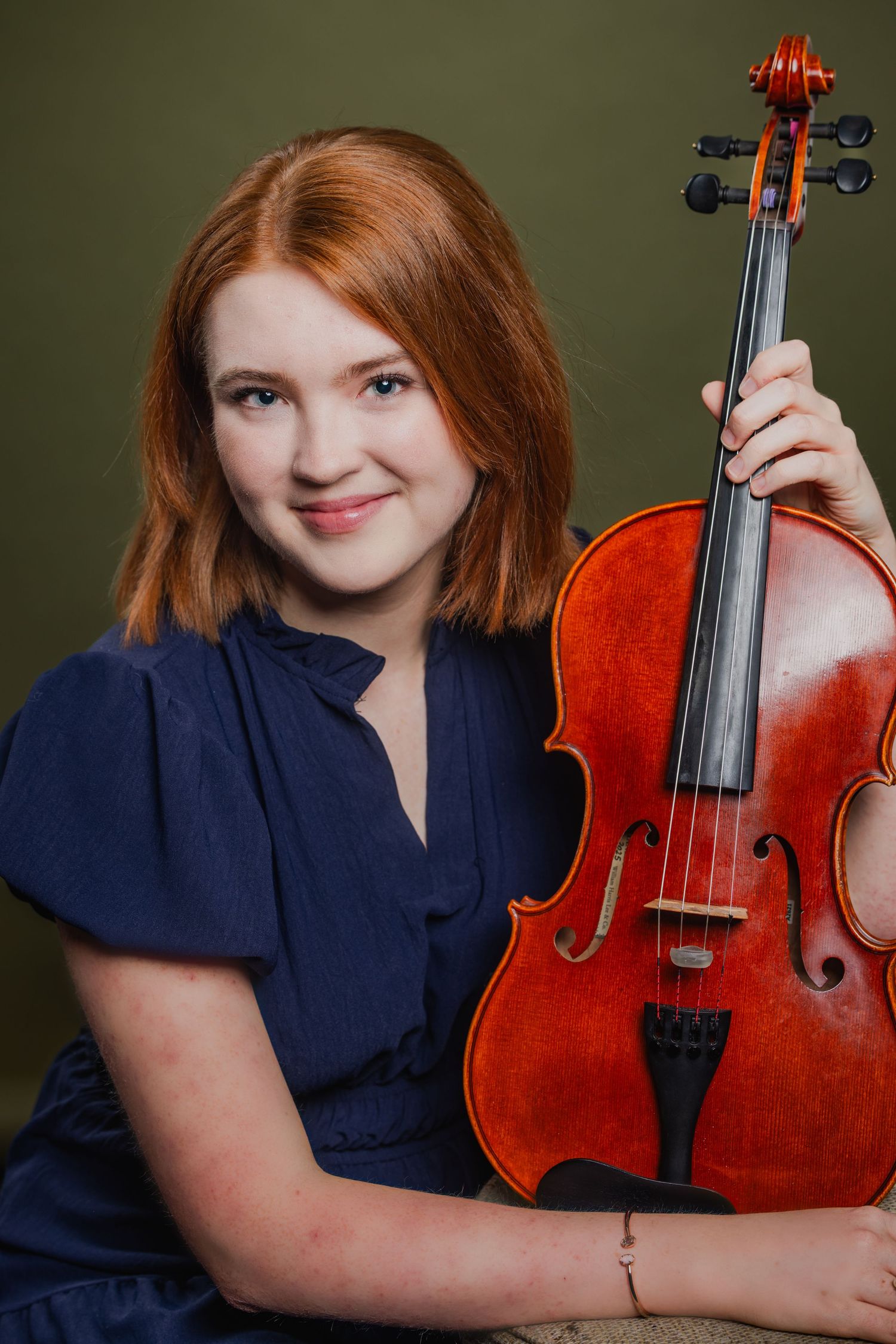 Young woman with red hair holding a cello, wearing a navy blue dress against an olive green background.