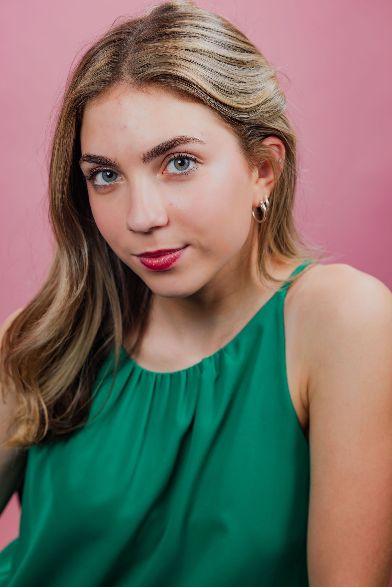 Young blonde woman in a green sleeveless top with pink lipstick against a pink background.
