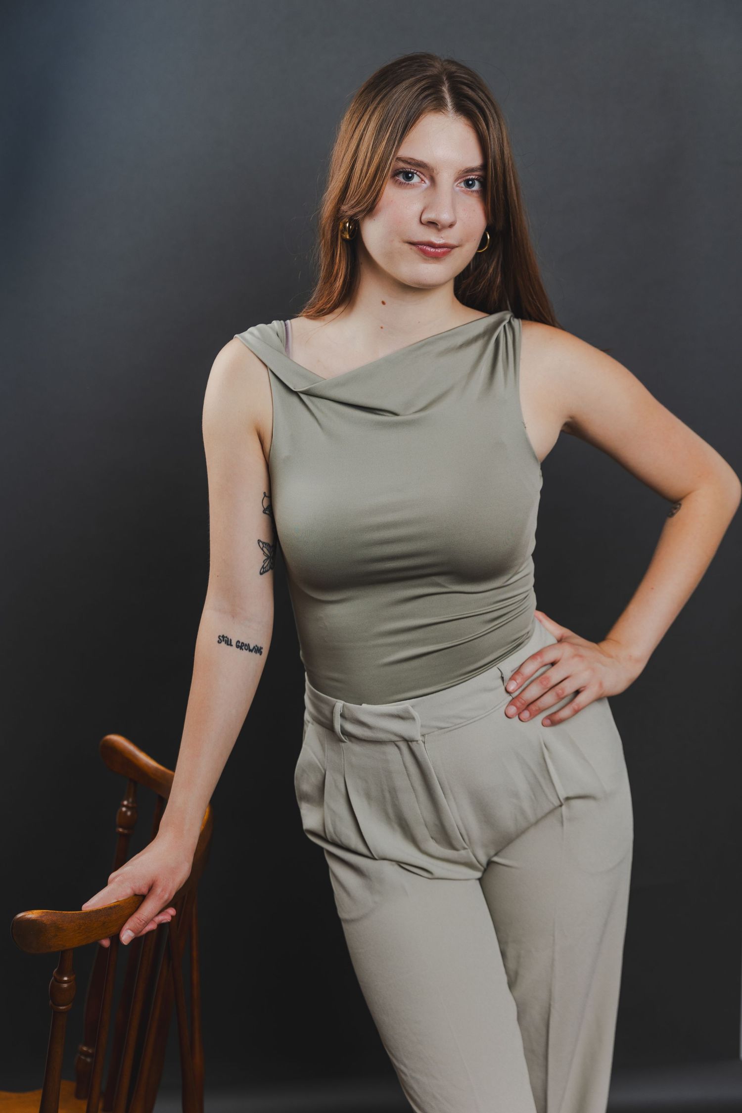 Young woman in a sage green sleeveless outfit poses beside a wooden chair against a gray background.