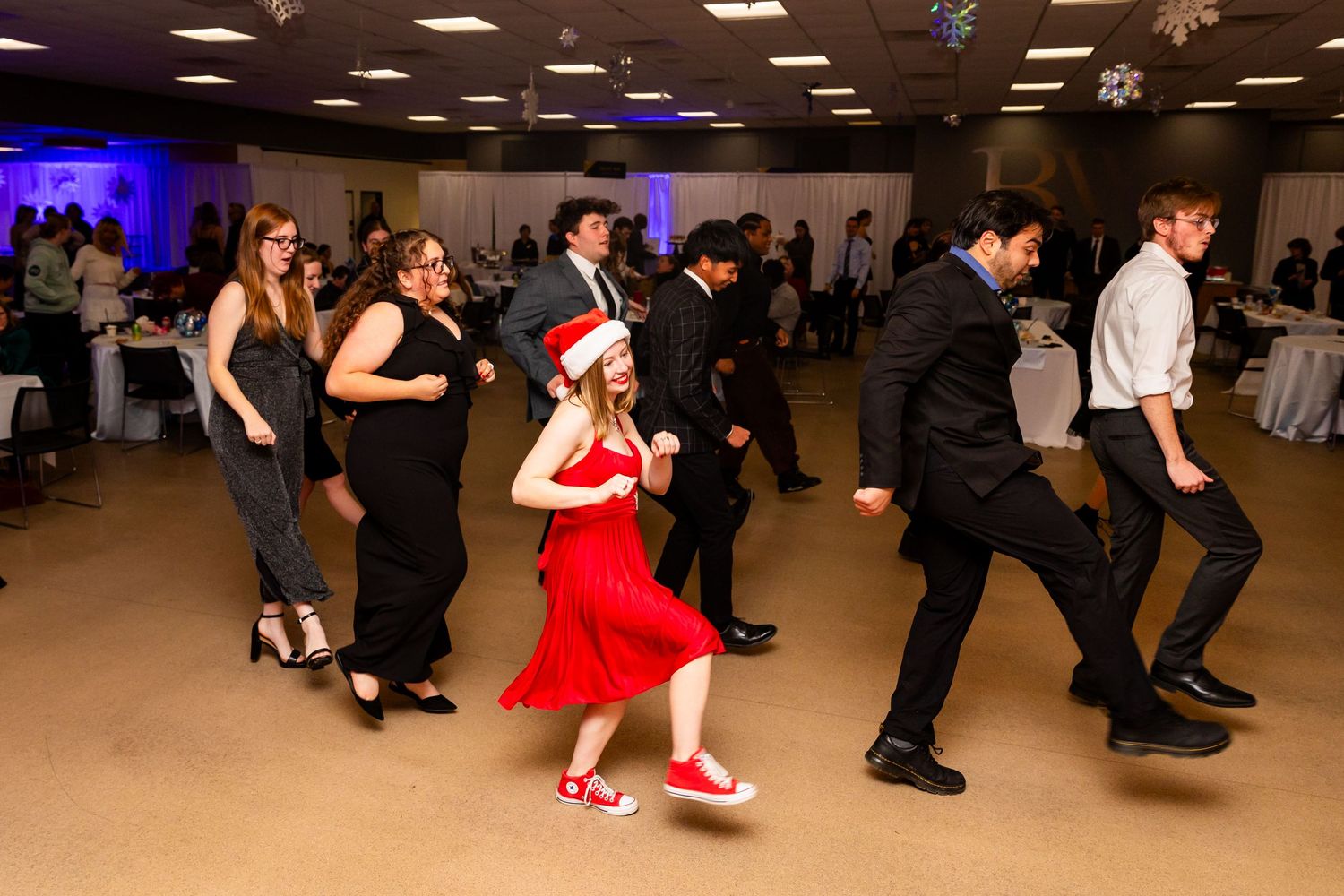 Students dancing at a festive holiday party with a girl in red dress and Santa hat leading the fun.