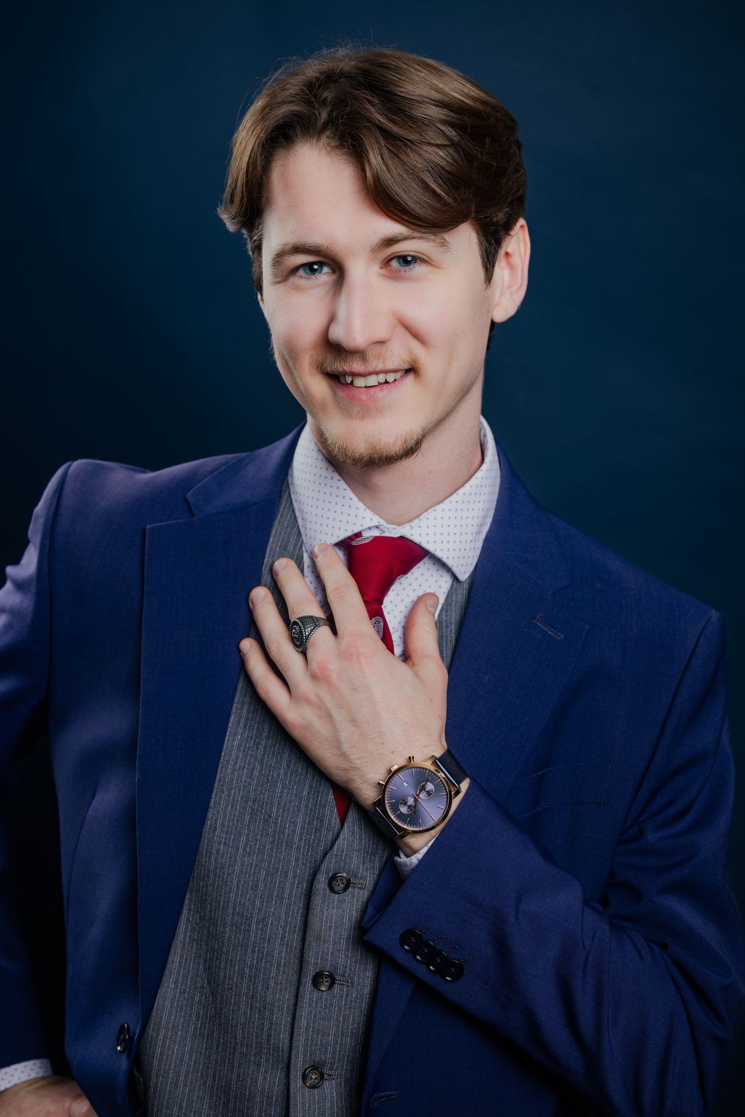Smiling young man in a blue suit with red tie and vest posing against a dark teal studio background.