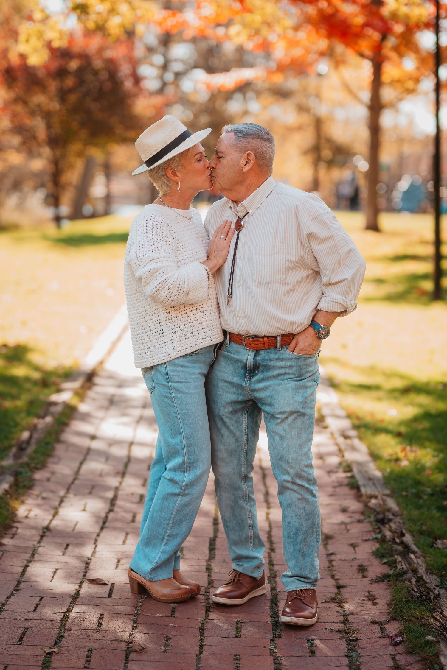 Senior couple sharing a romantic kiss on brick pathway surrounded by colorful autumn trees.