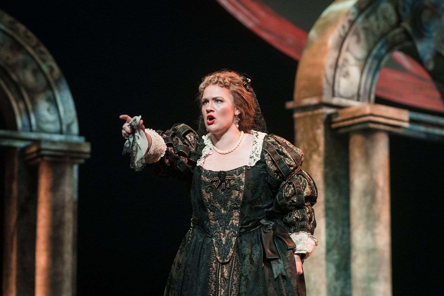 Female opera singer in a dark green Renaissance costume gesturing dramatically under stage lighting.