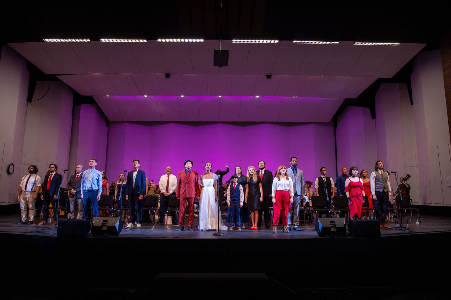 A diverse ensemble of performers takes a bow on a purple-lit concert stage.