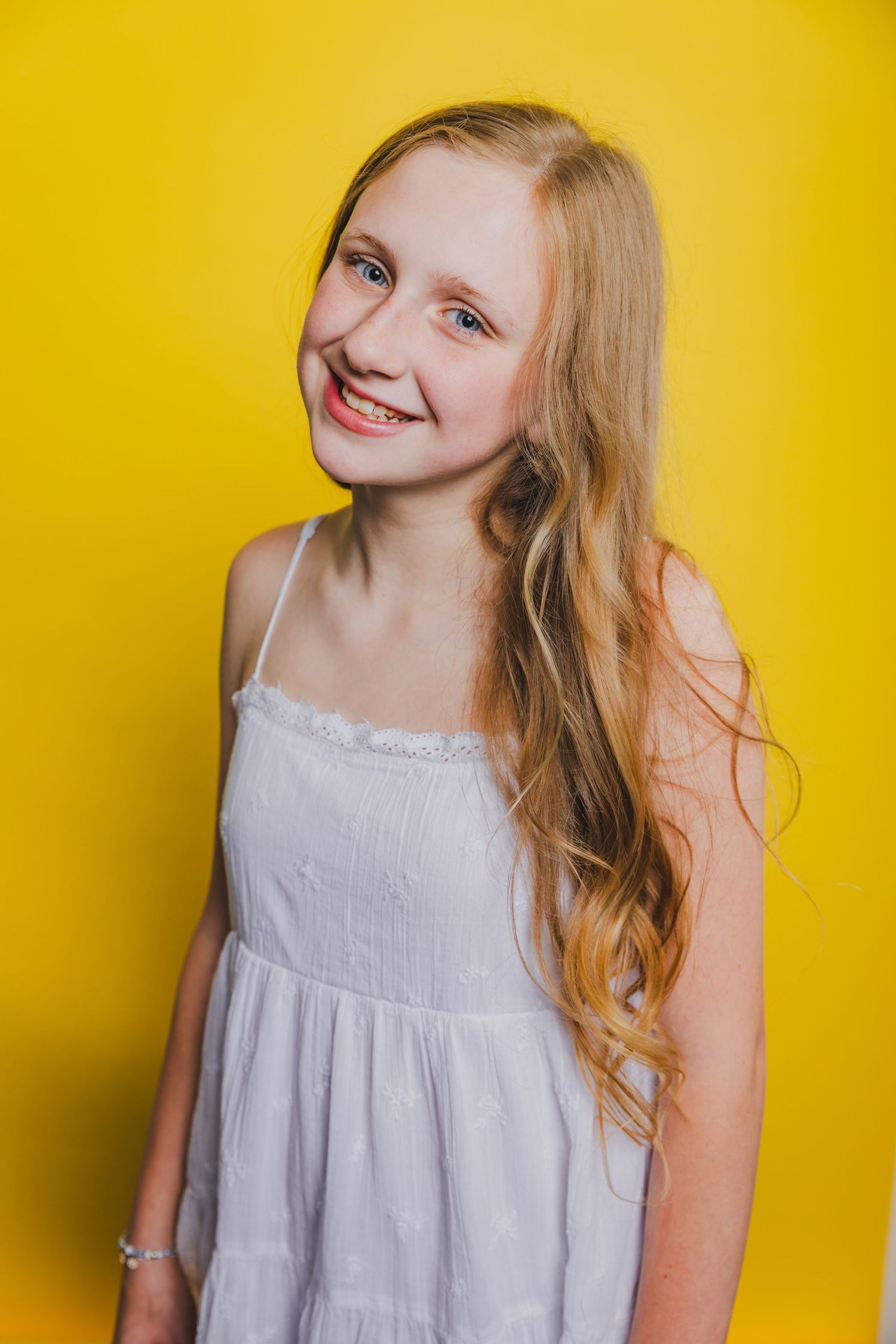 Blonde teenage girl with long wavy hair in a white dress smiling against a yellow background.