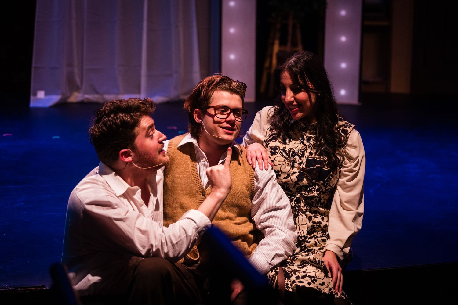 Three young theater performers share an intimate scene under blue stage lighting in a musical production.